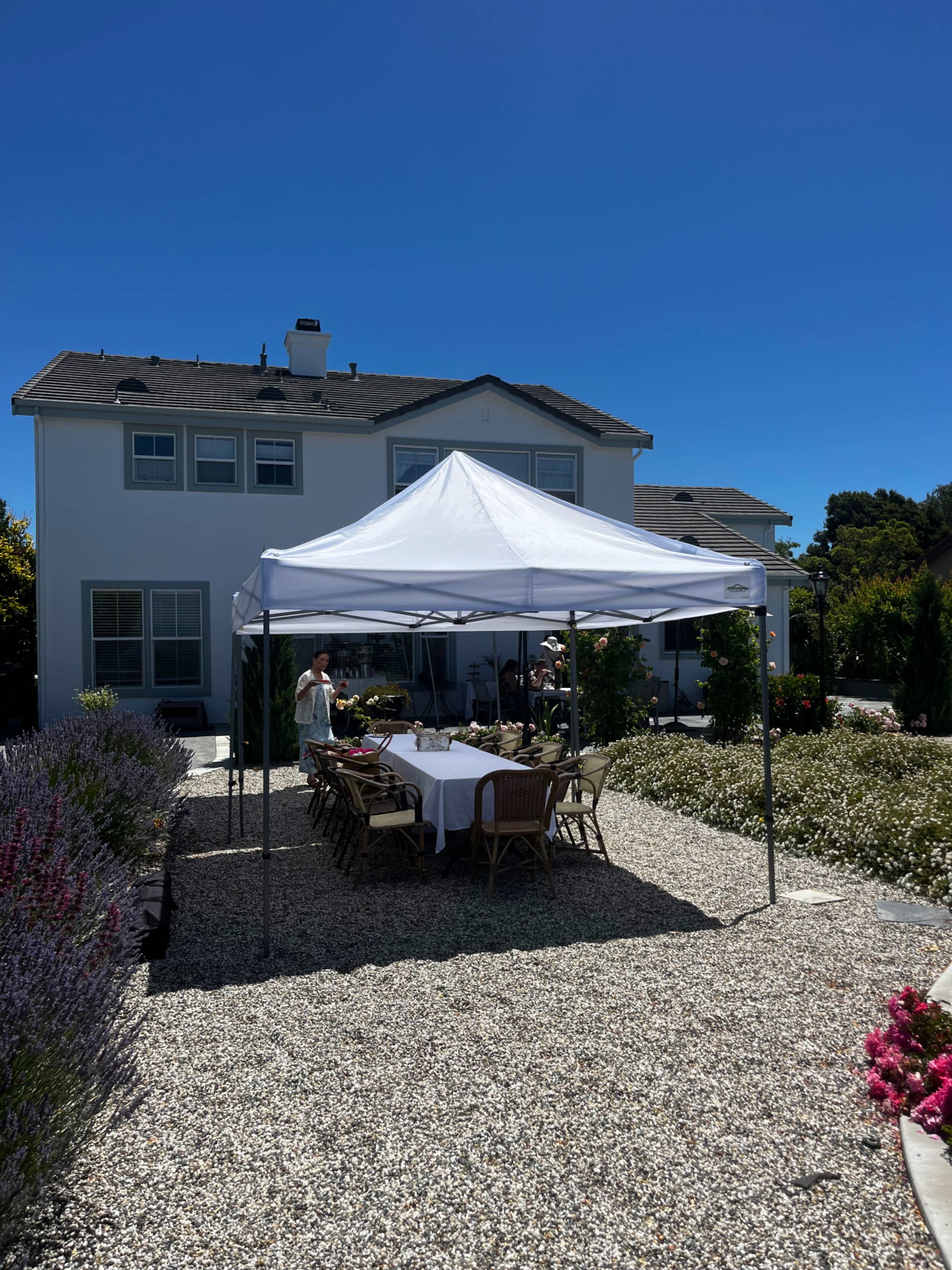 A white canopy tent is set up in a gravel area with tables and chairs arranged beneath it, in front of a large white house surrounded by colorful flowers.