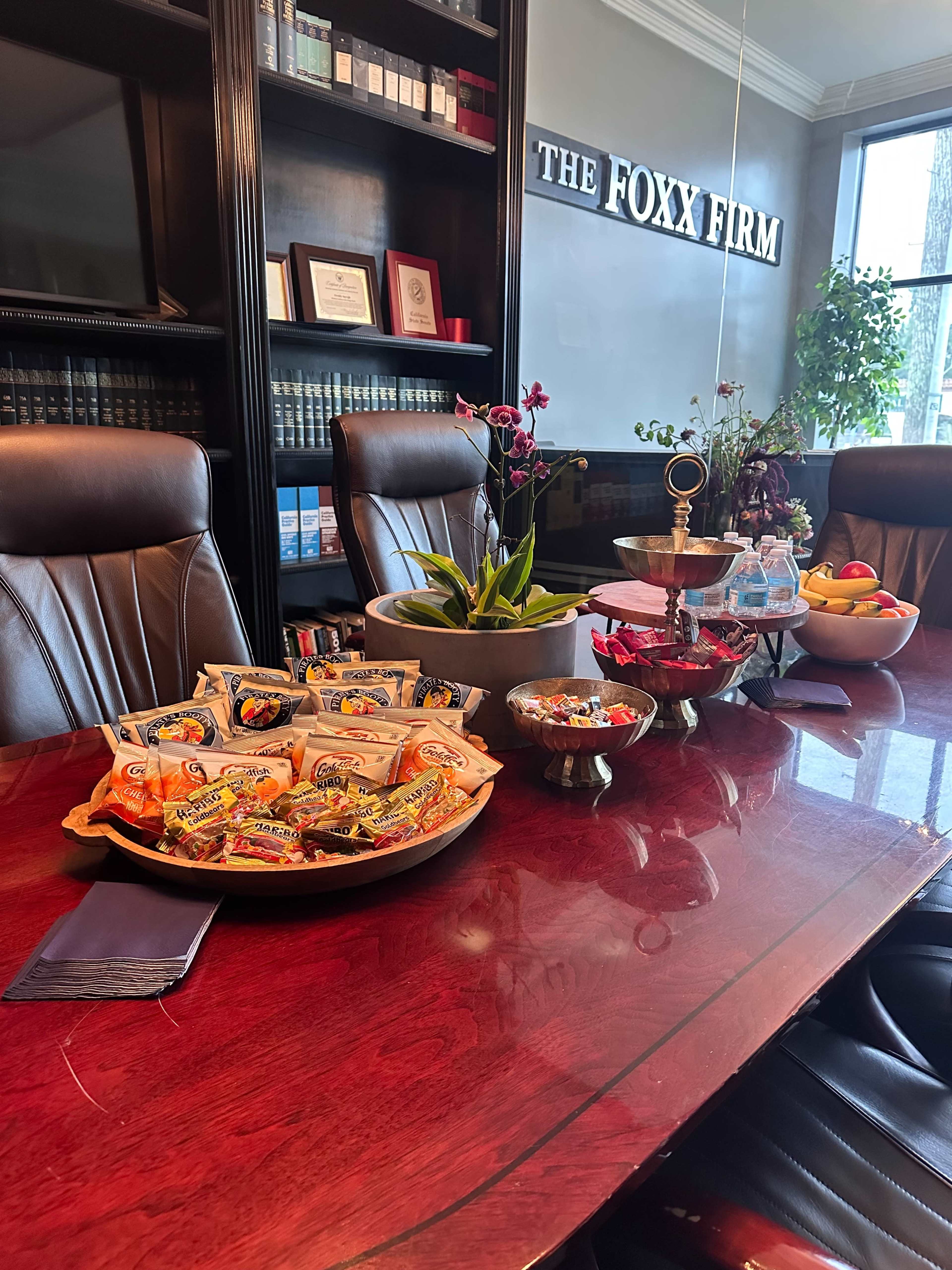 The image shows a conference table in an office setting, adorned with a variety of snacks, fruits, and decorative plants, alongside leather chairs and shelves filled with books.