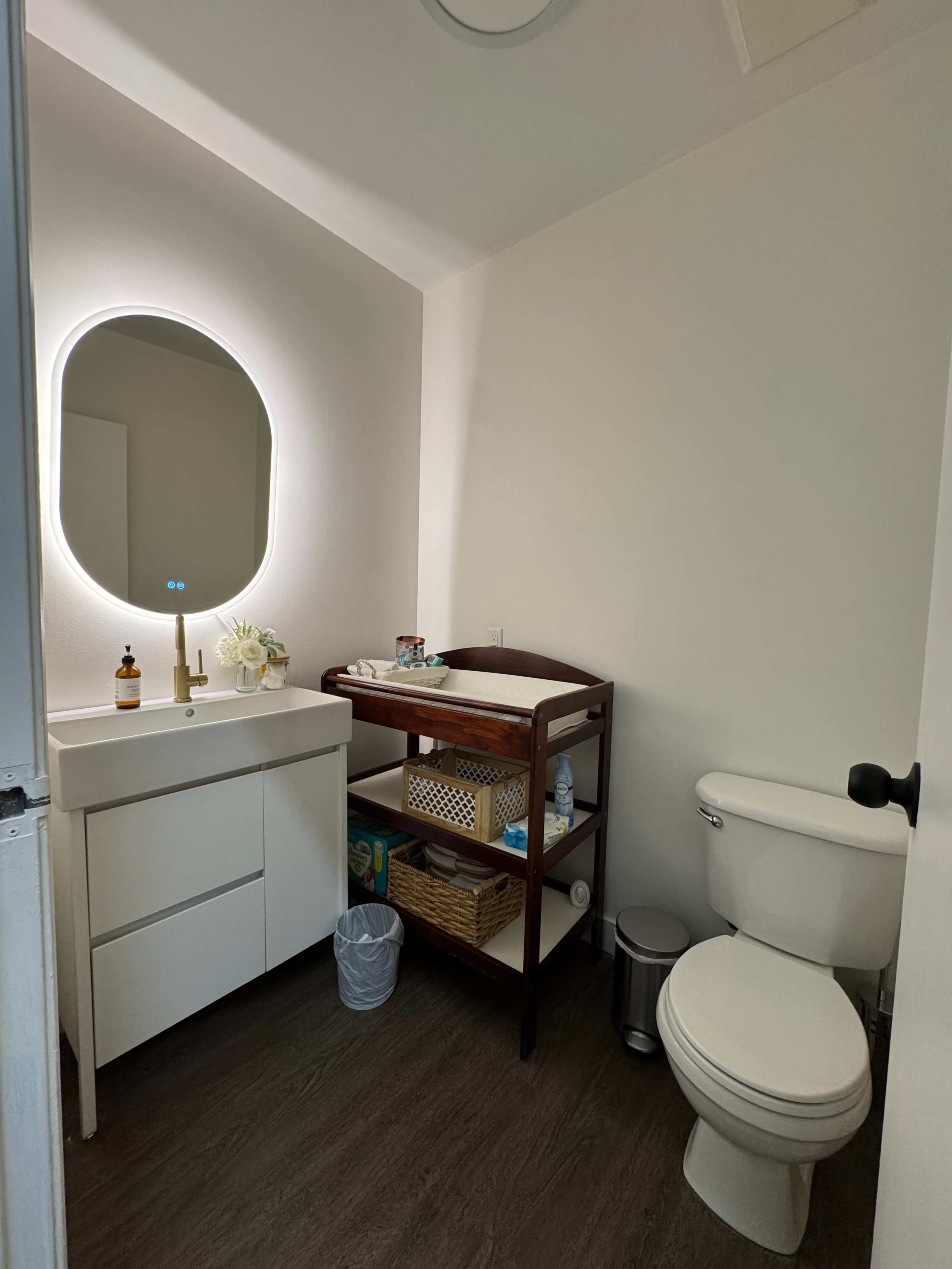The image shows a small bathroom with a toilet, a wooden cart holding supplies, and a mirror with ambient lighting.