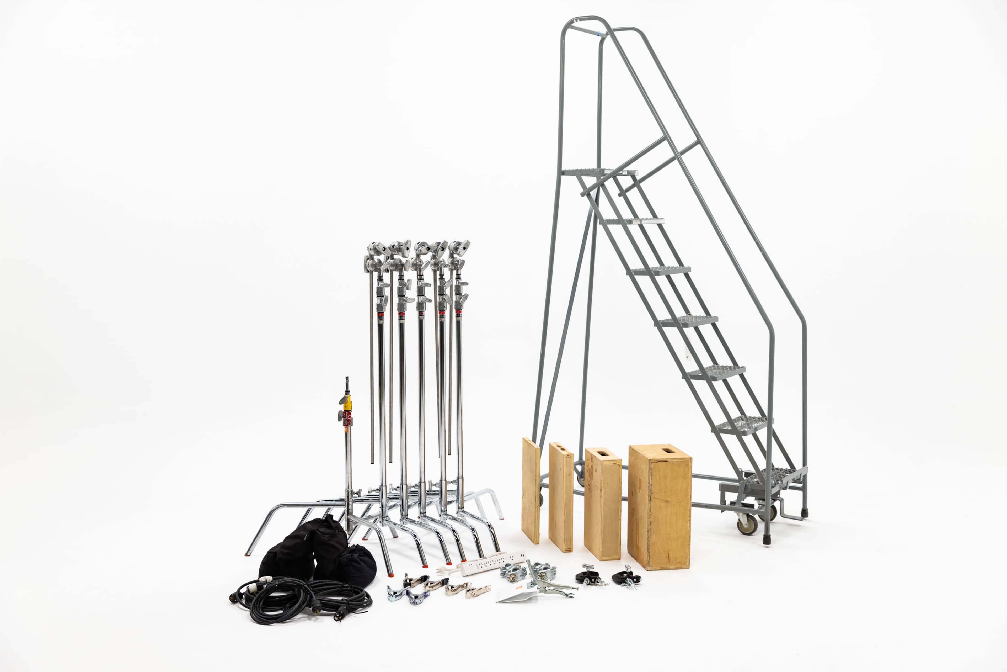 The image shows a collection of grip equipment including several light stands, a wooden apple box, and a set of stairs against a white background.