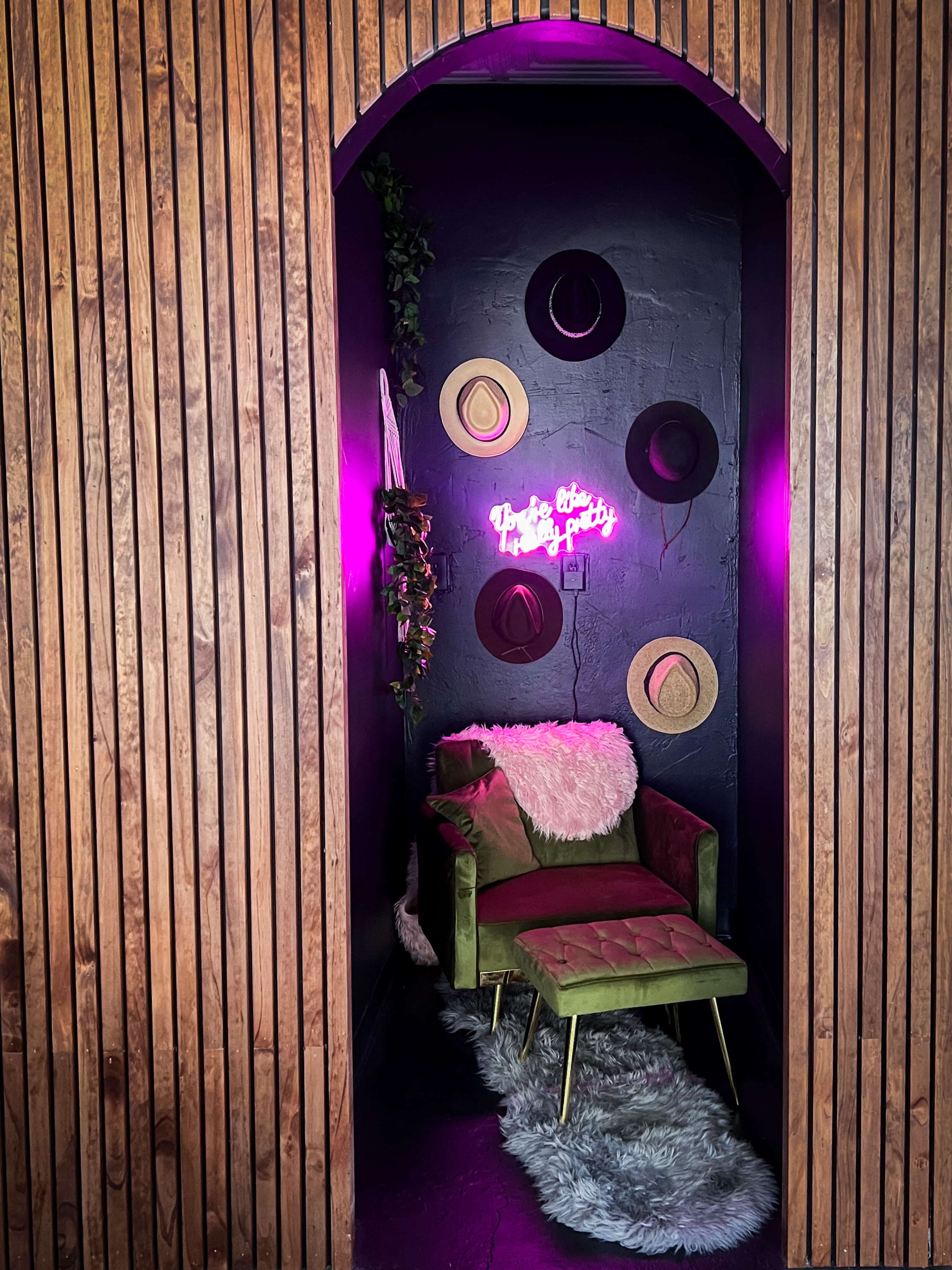 A green chair sits on a textured rug in an alcove with wooden walls, adorned with decorative hats and a neon sign.