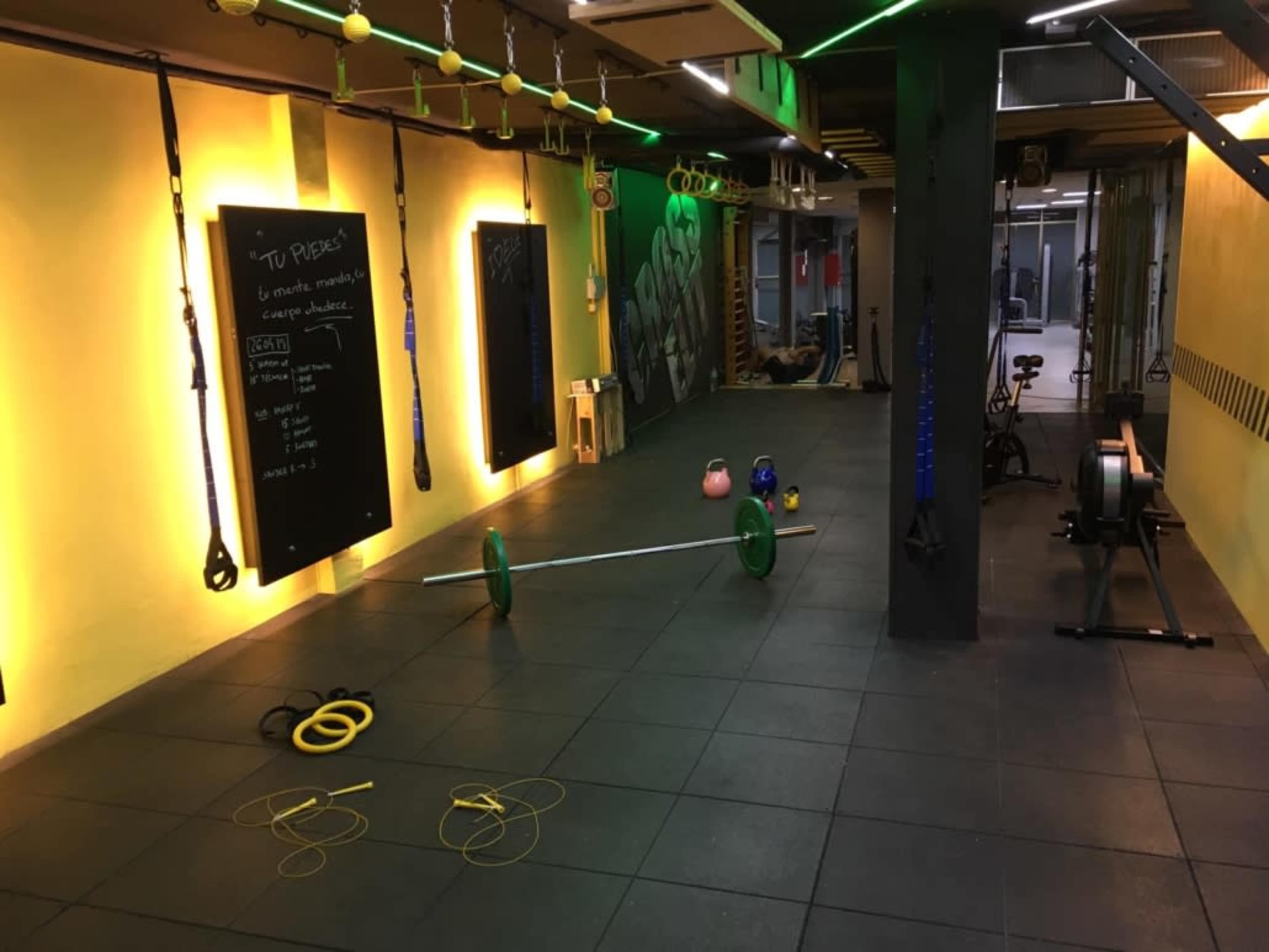 The interior of a gym features various exercise equipment, including barbells, kettlebells, and suspension trainers, set on a black rubber floor with yellow walls.