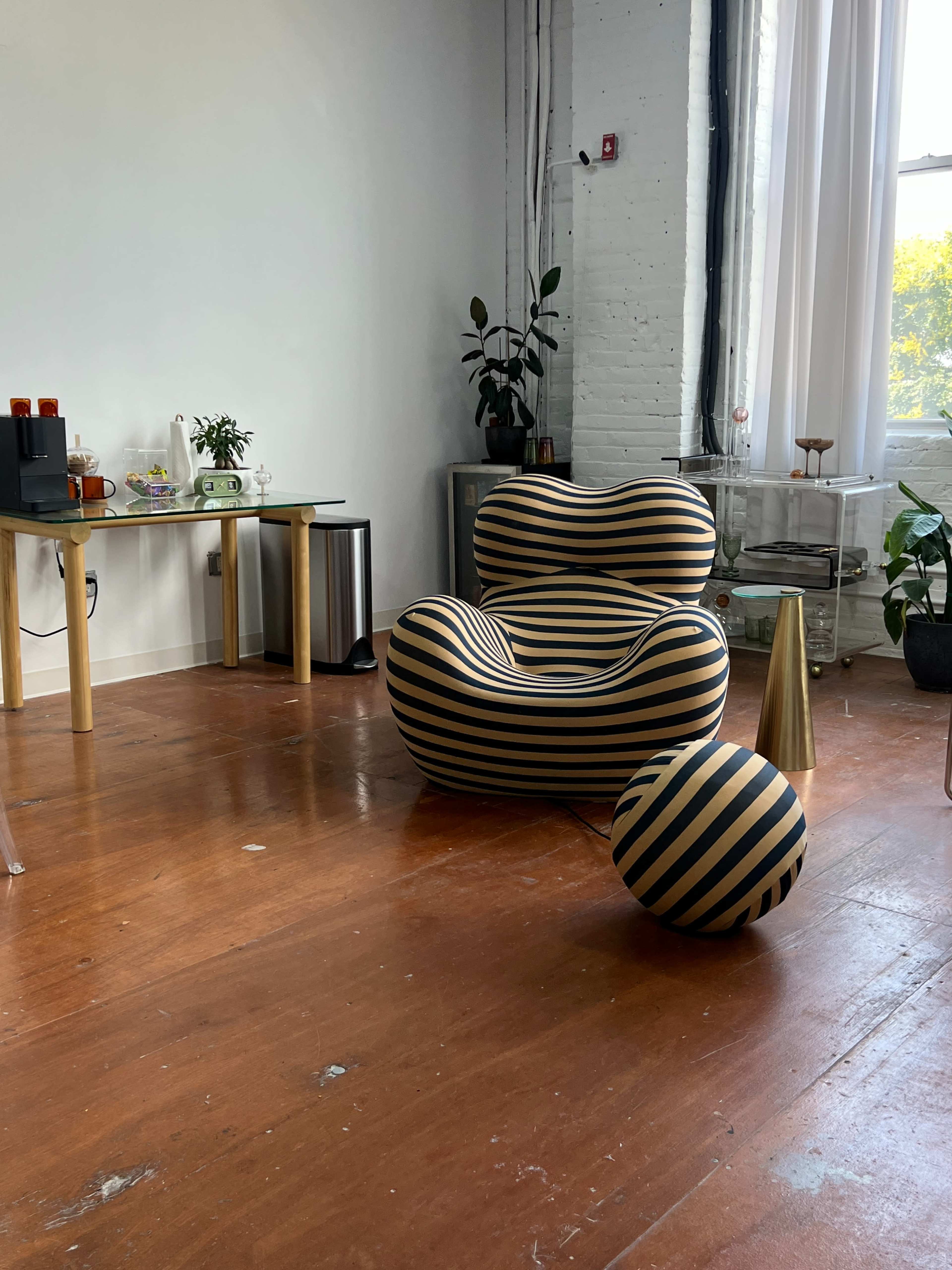The image shows a modern, striped chair accompanied by a matching round ball in a bright room with wooden flooring and plants.