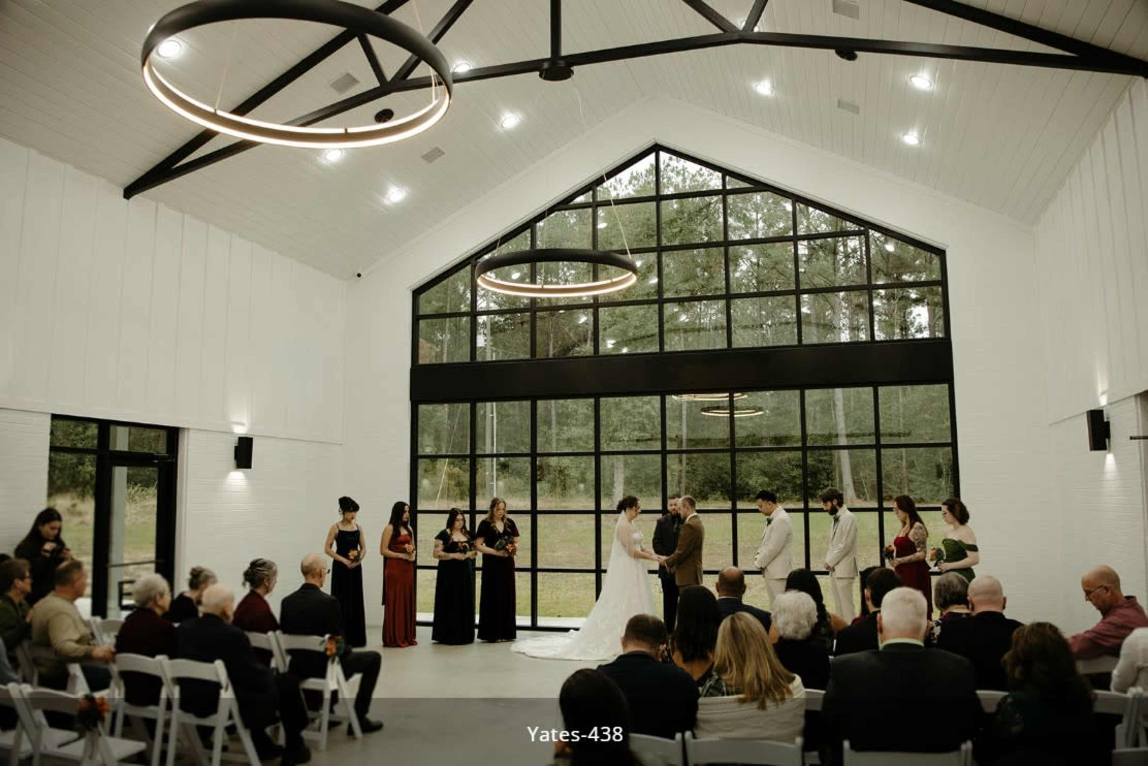 A wedding ceremony takes place in a modern venue with large windows, where the couple stands at the altar surrounded by their wedding party and guests seated nearby.
