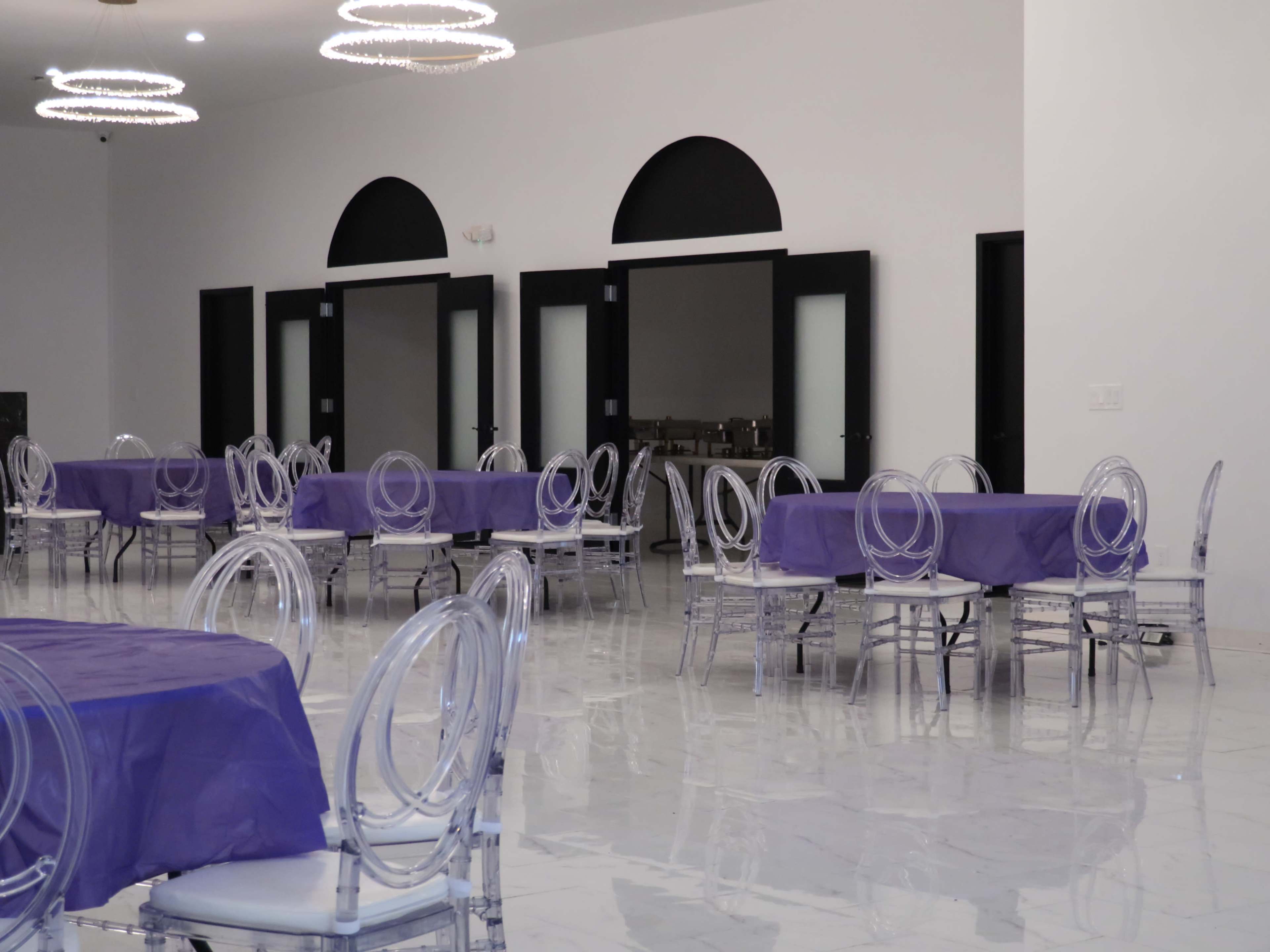 A spacious event hall with several tables covered in purple tablecloths and transparent chairs arranged on a glossy white floor.