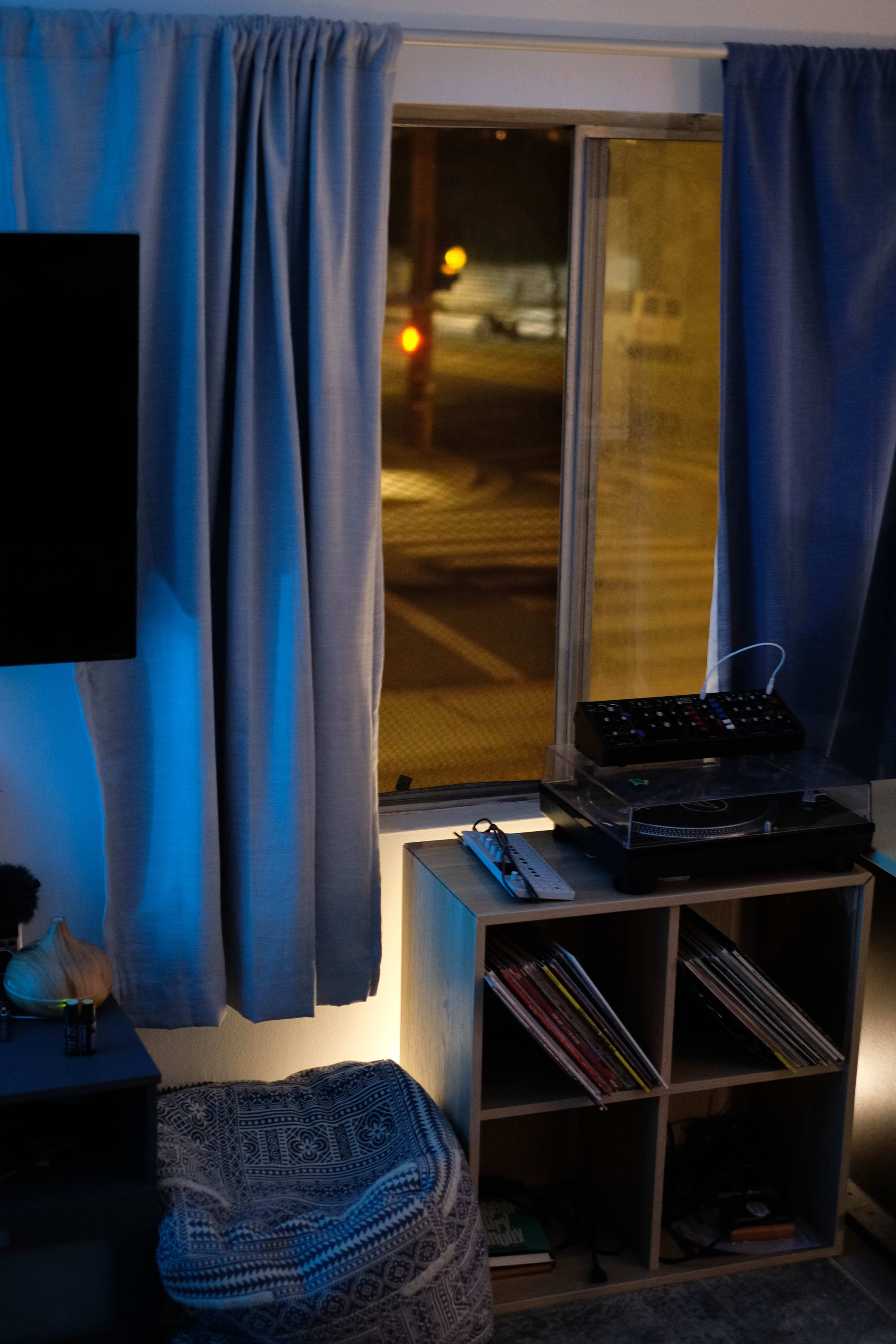 A cozy room features a window with curtains, a record player on a shelving unit, and a stack of vinyl records beside a textured ottoman.