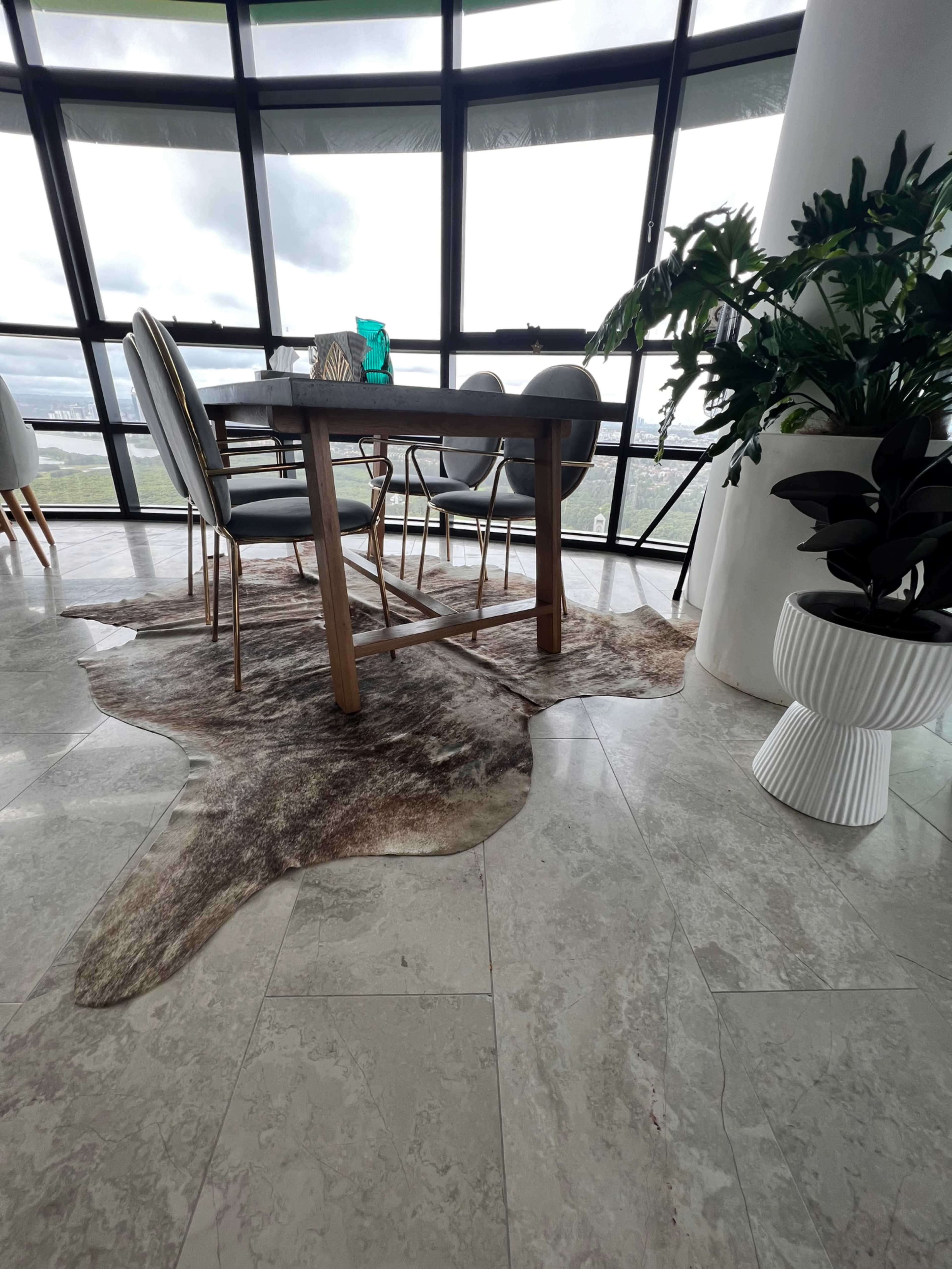 The image shows a modern dining area with a wooden table and chairs placed on a cowhide rug, surrounded by large windows that provide a view of the outside.