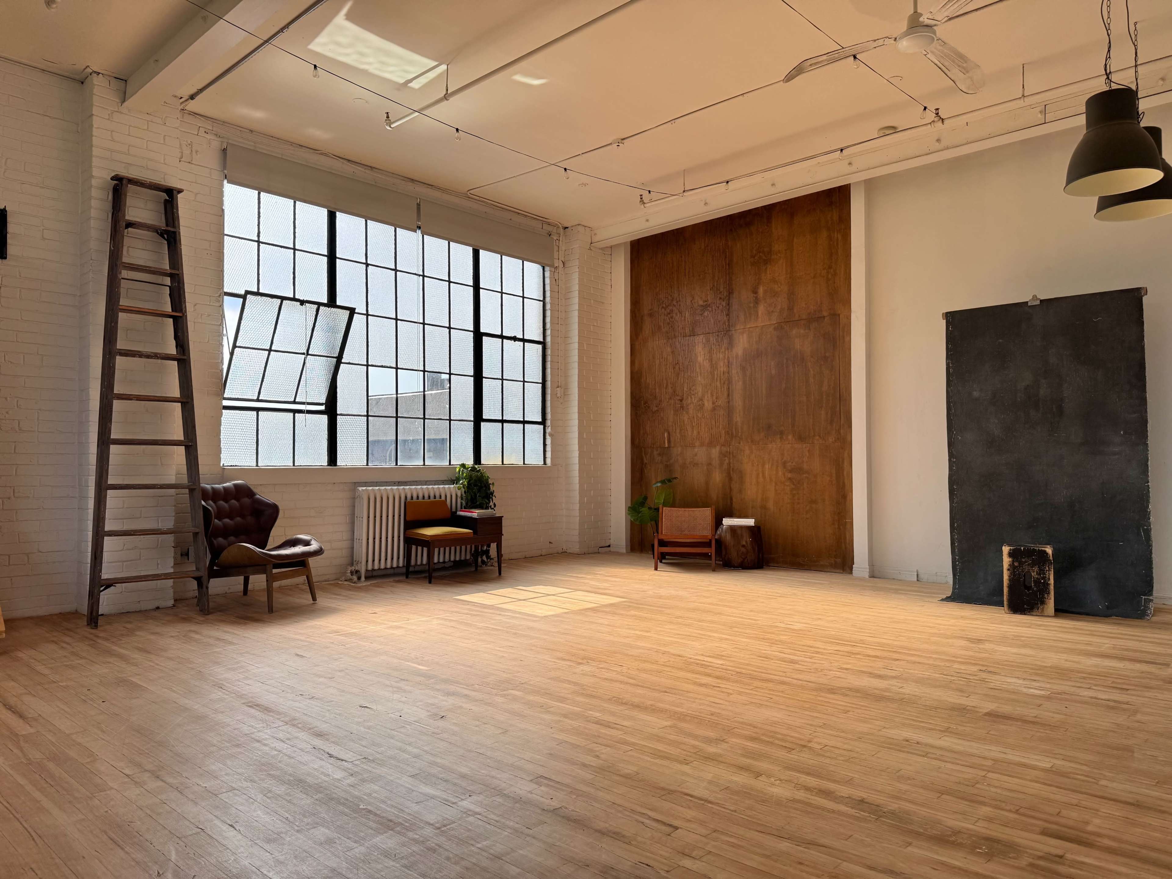 The image depicts a spacious, well-lit studio with a wooden floor, large windows, a ladder, and minimal furnishings including a chair and a small table.