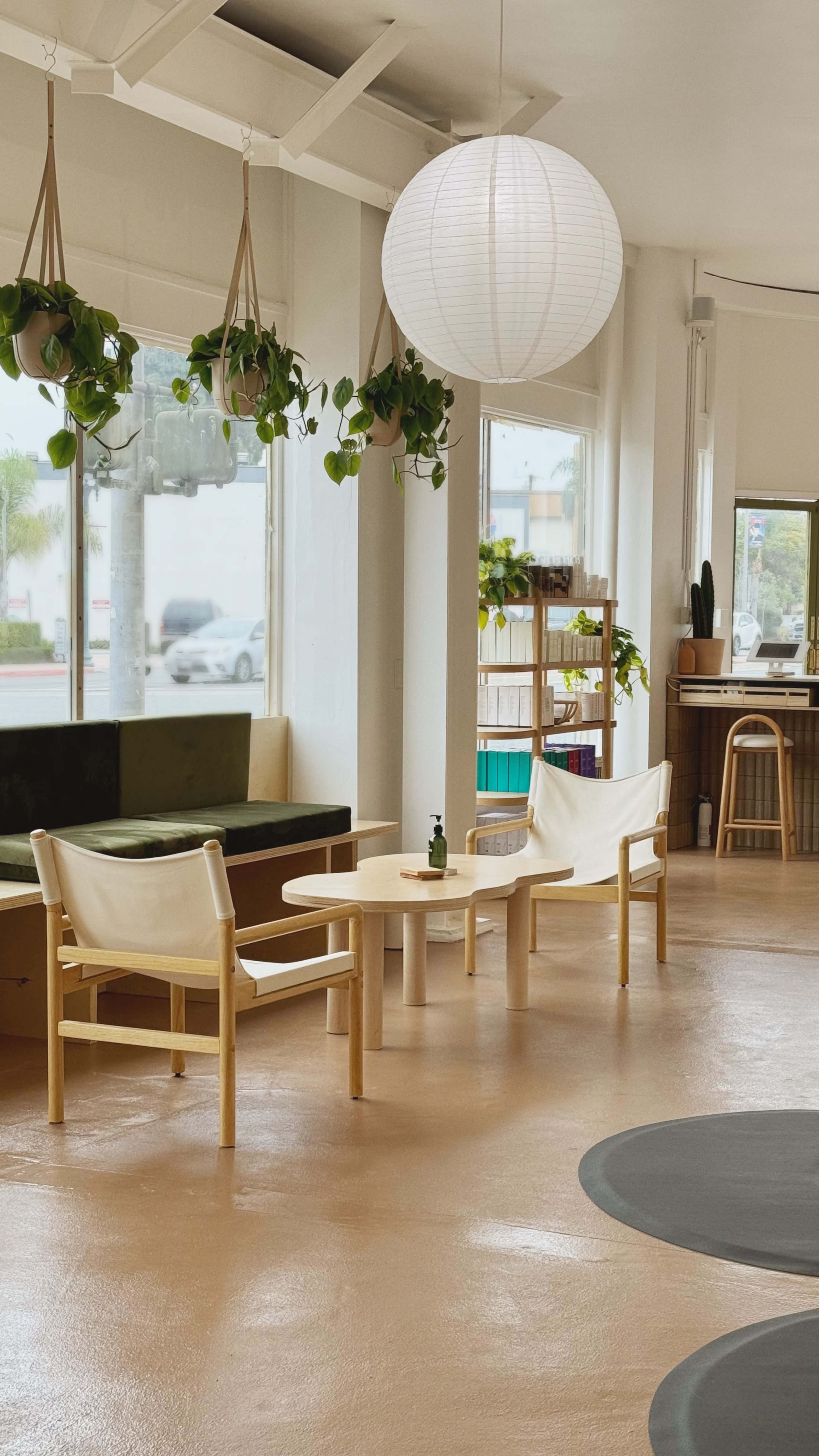 A light-filled café features two minimalist chairs and a round table surrounded by hanging plants and a large paper lantern.