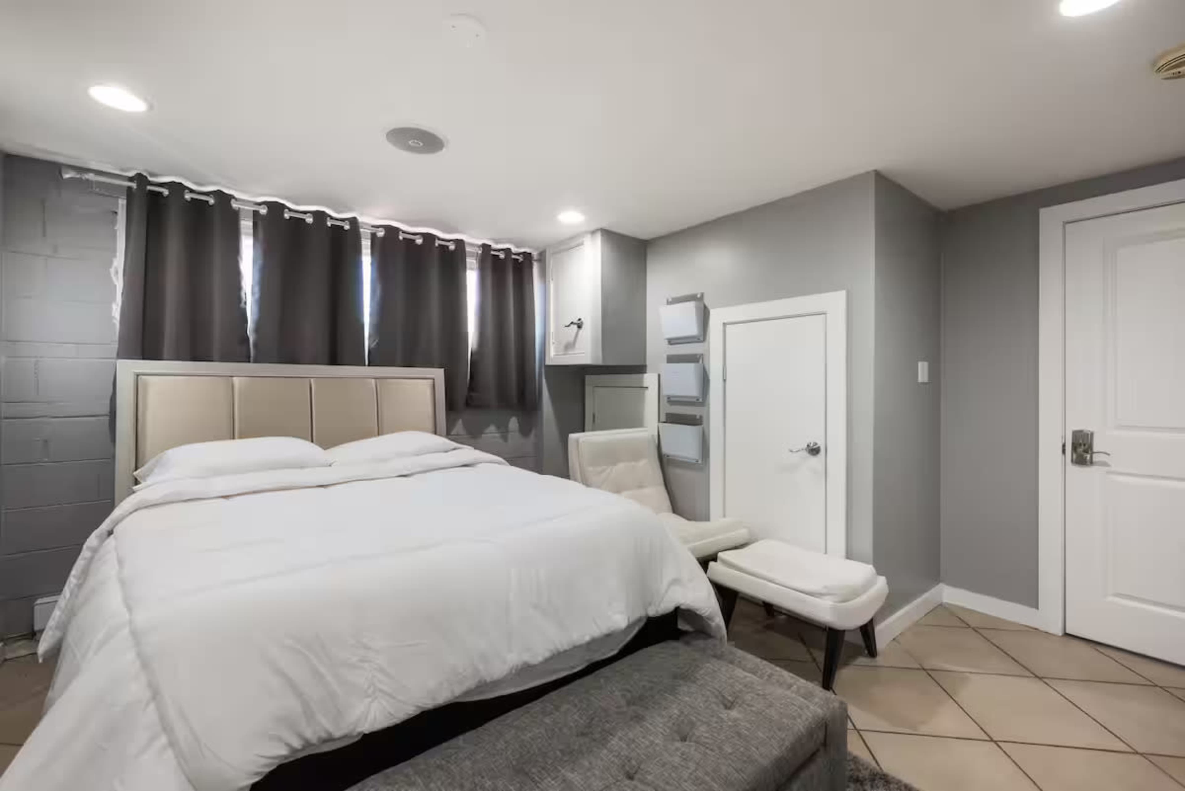 The image shows a modern bedroom with a bed, grey walls, and a small seating area.