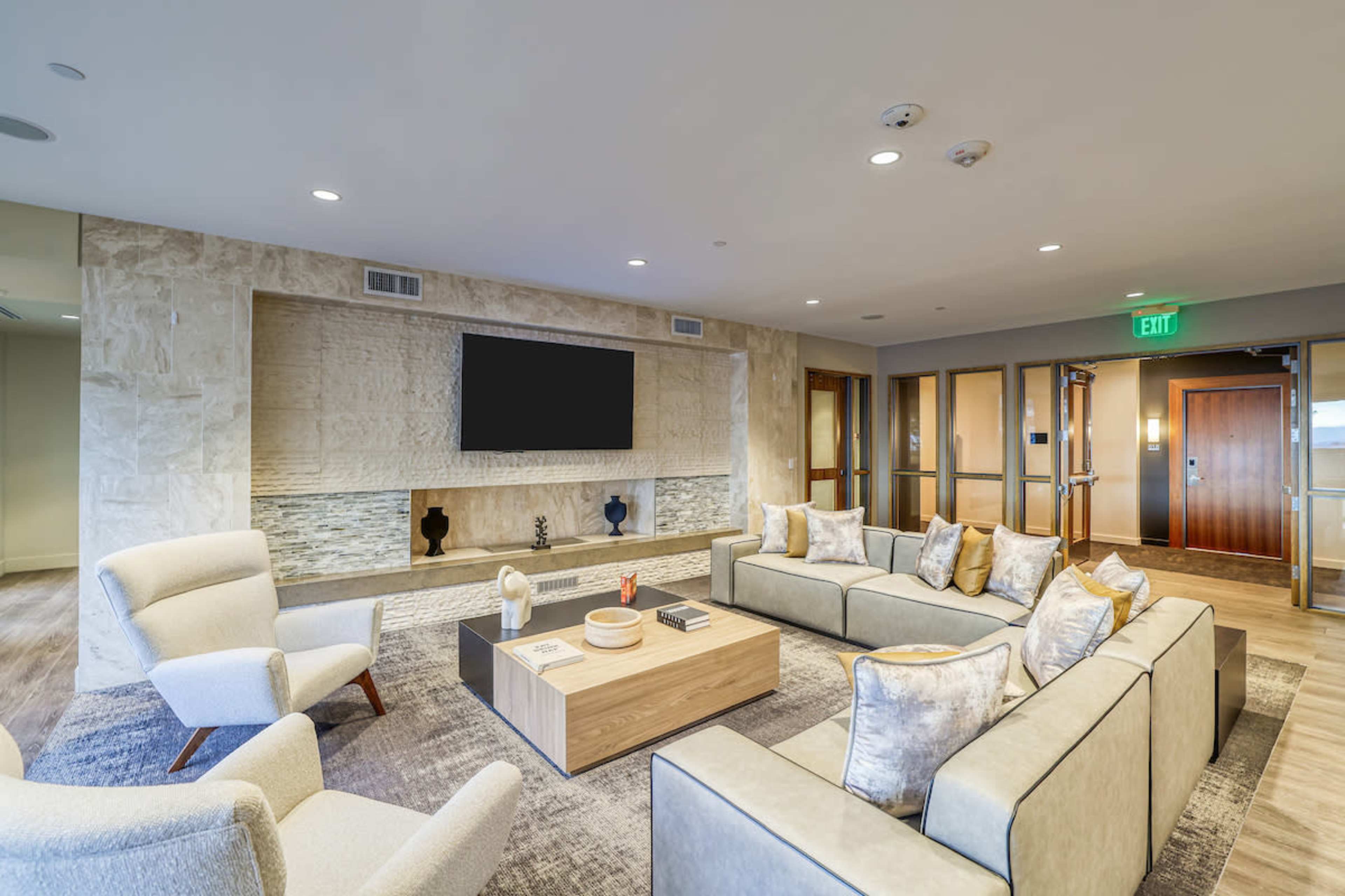 The image shows a modern lounge area featuring a large flat-screen television mounted above a stone accent wall, with a sectional sofa and armchairs arranged around a central coffee table.