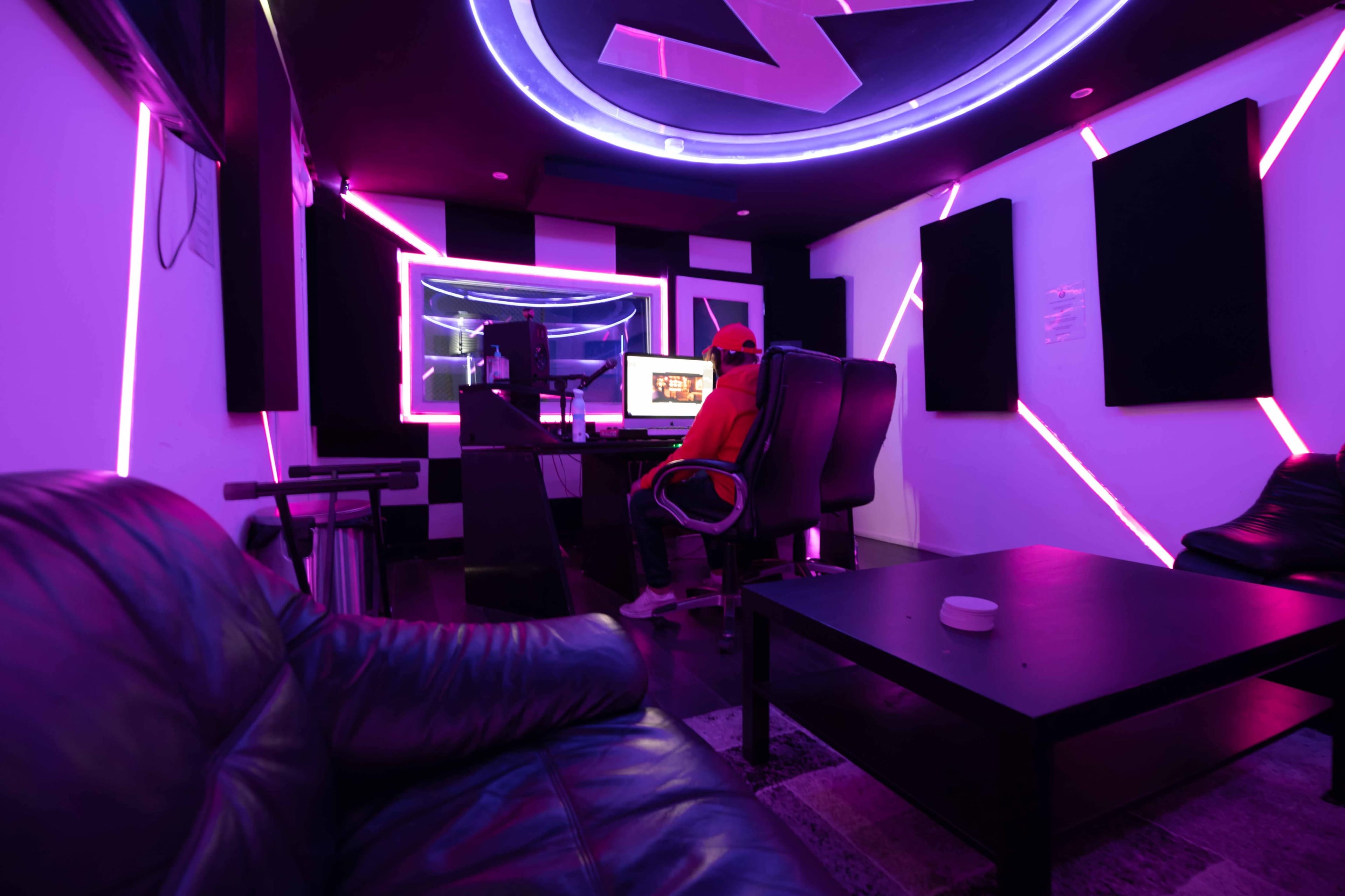 A person in a red hoodie sits at a desk in a music studio, surrounded by purple-lit walls and a black leather couch.