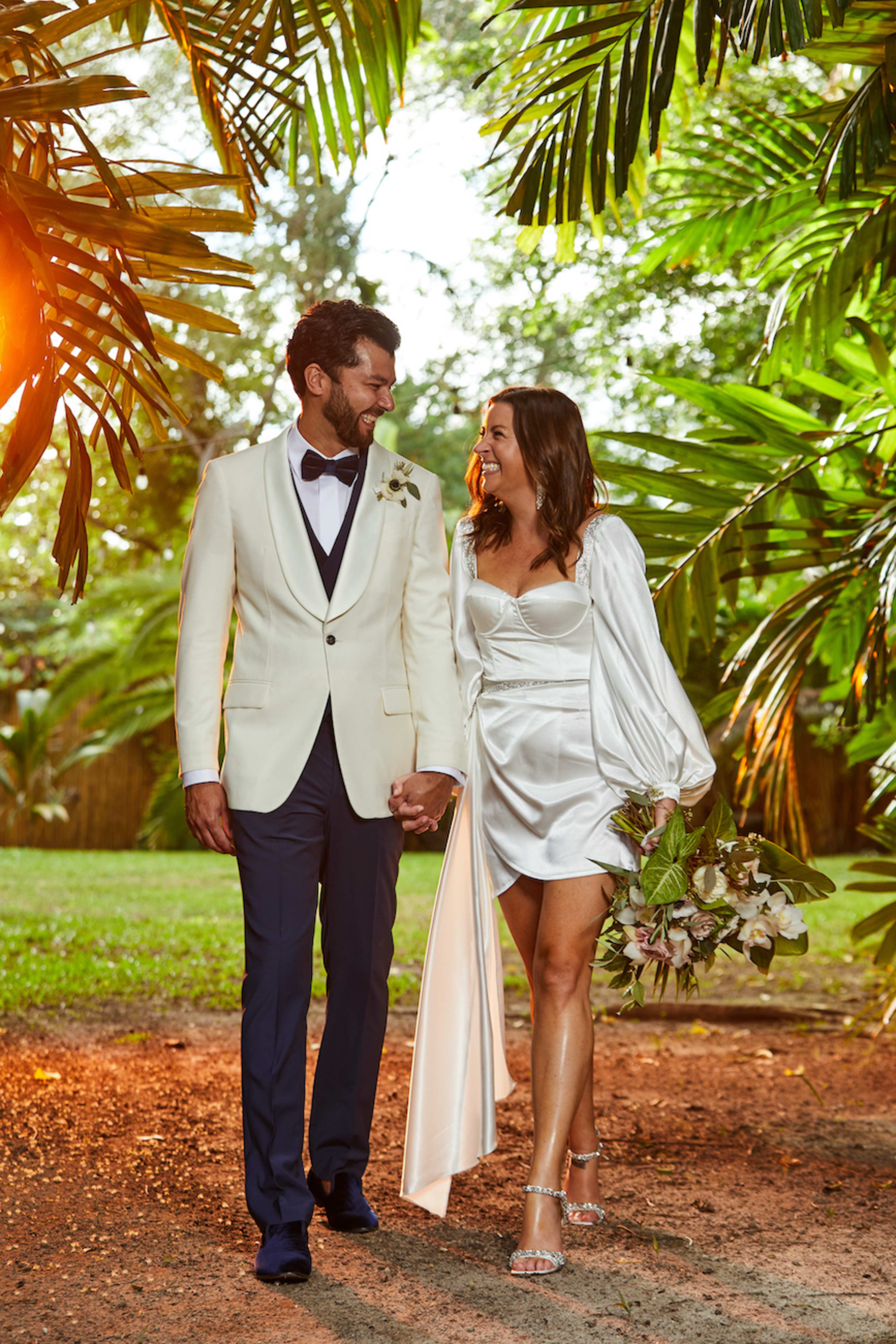 A couple in formal attire walks hand-in-hand through a lush outdoor setting, surrounded by palm leaves.