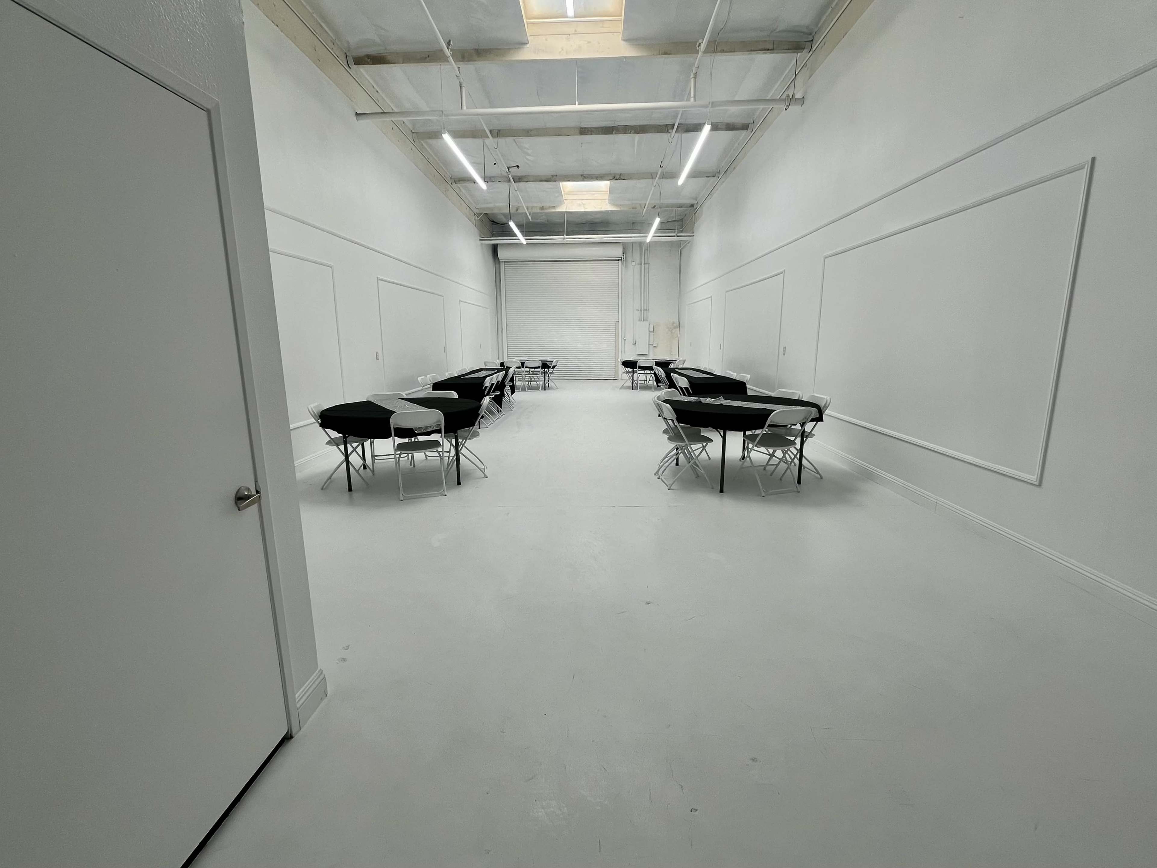 The image shows a large, empty room with white walls, a smooth white floor, and several black folding tables arranged in two rows.