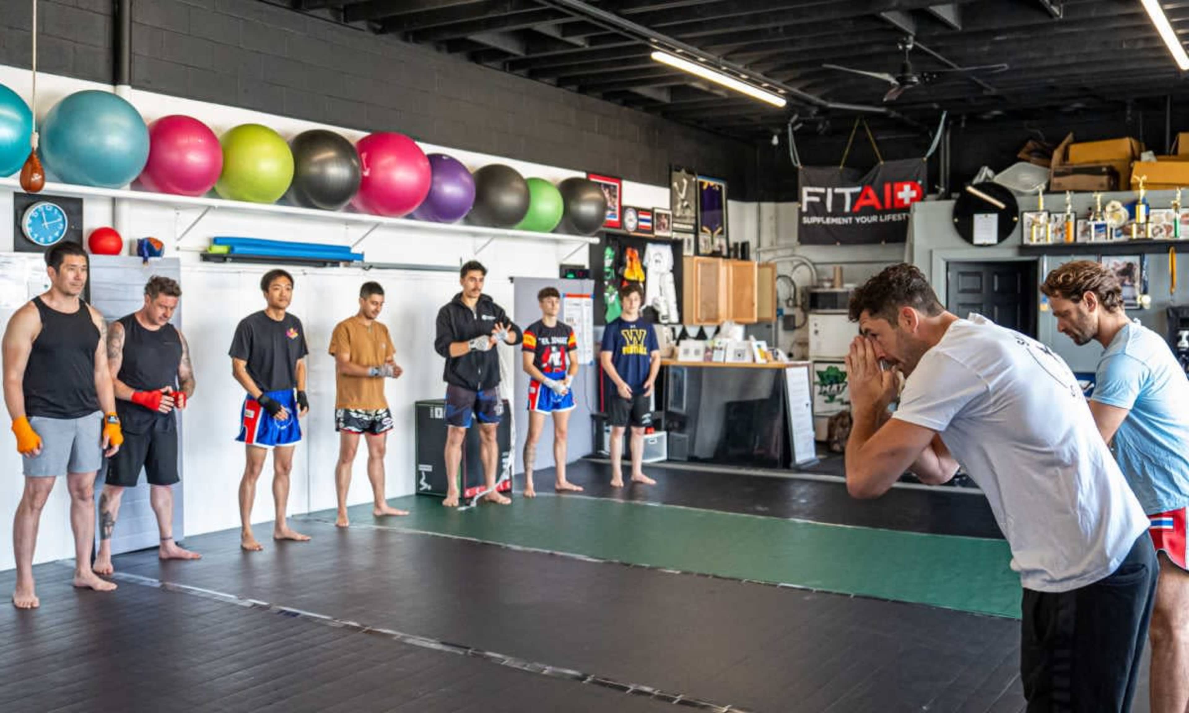 A group of eight people stands in a gym, preparing for a martial arts training session, while two individuals demonstrate techniques in the foreground.