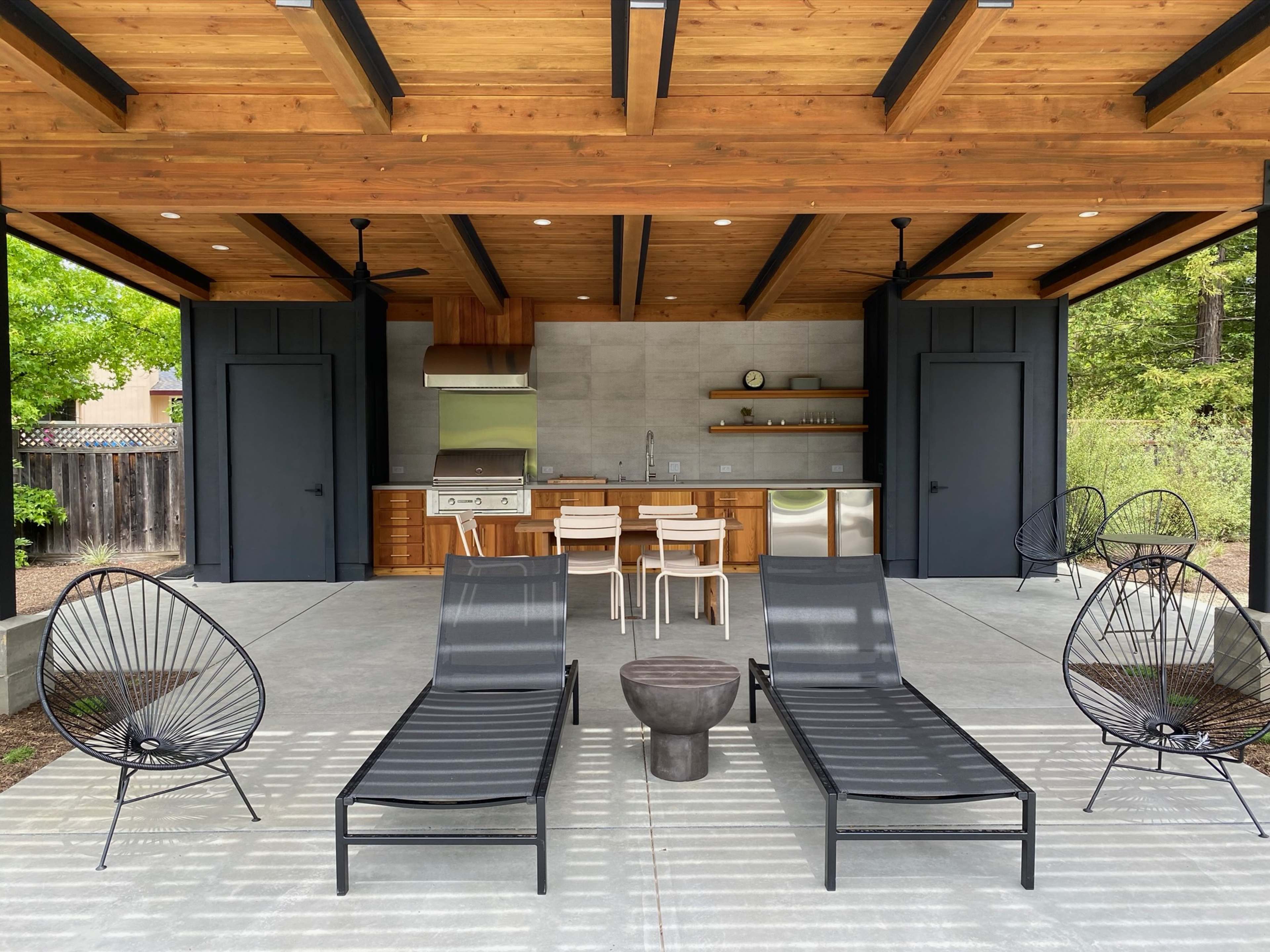 The image shows a modern outdoor patio with two black lounge chairs, a small round table, and a kitchen area featuring stainless steel appliances and wooden cabinetry under a wooden roof.