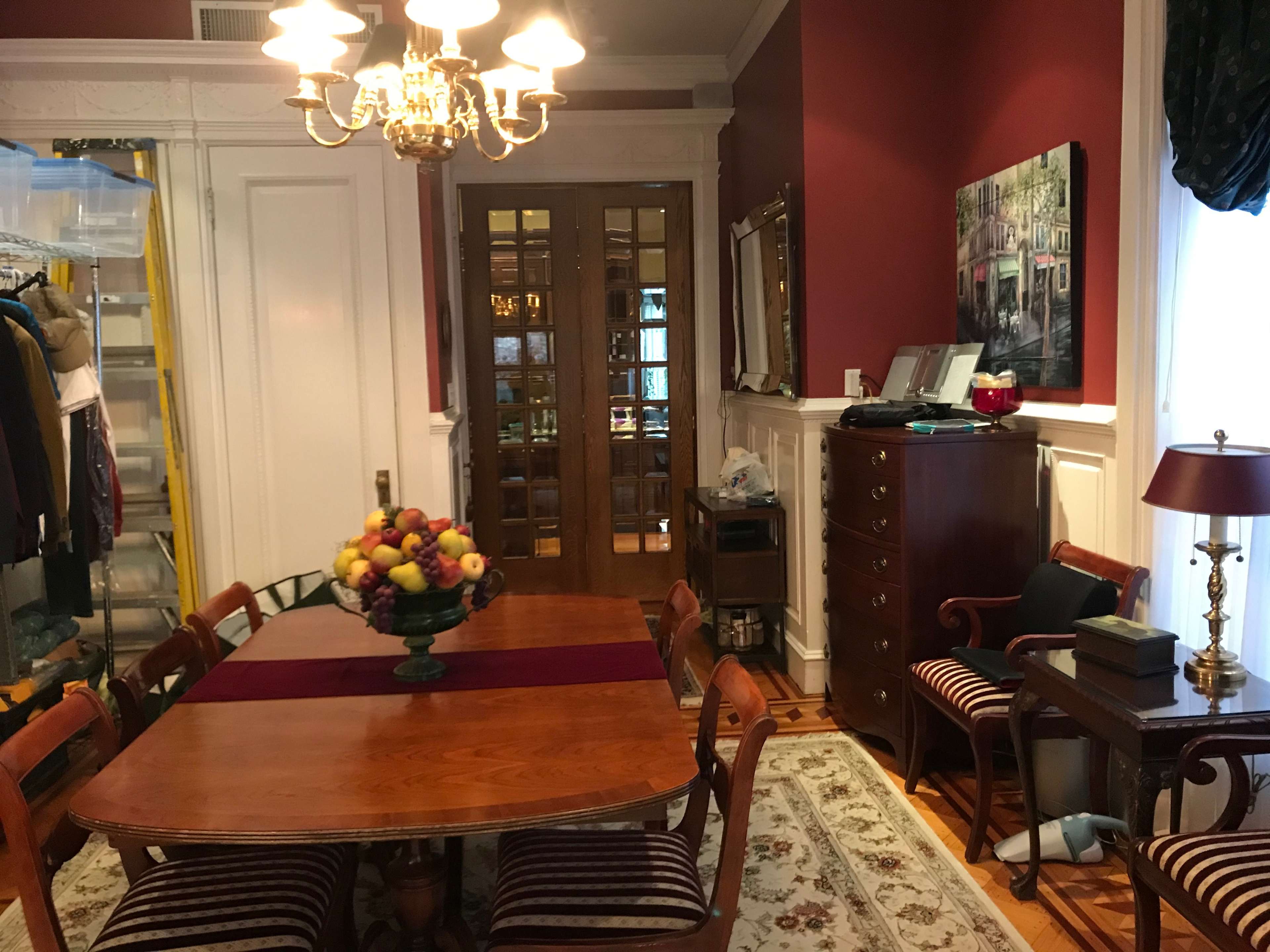 A dining room with a wooden table set for a meal, surrounded by chairs, and a large fruit bowl in the center, alongside a sideboard and a cabinet against the wall.