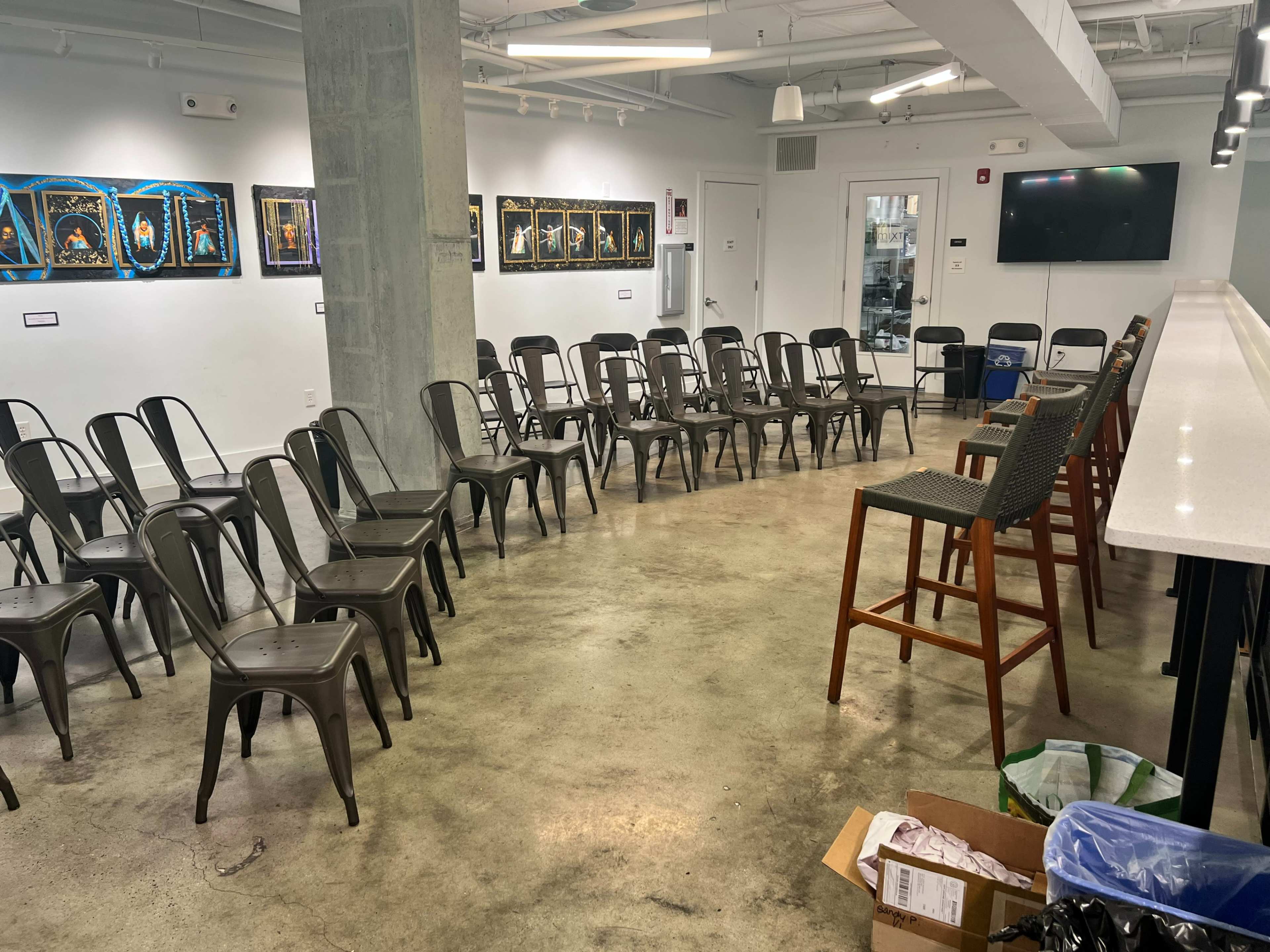 The image shows a room arranged with gray metal chairs in a circular formation around a central space, with a bar area and artwork displayed on the walls.