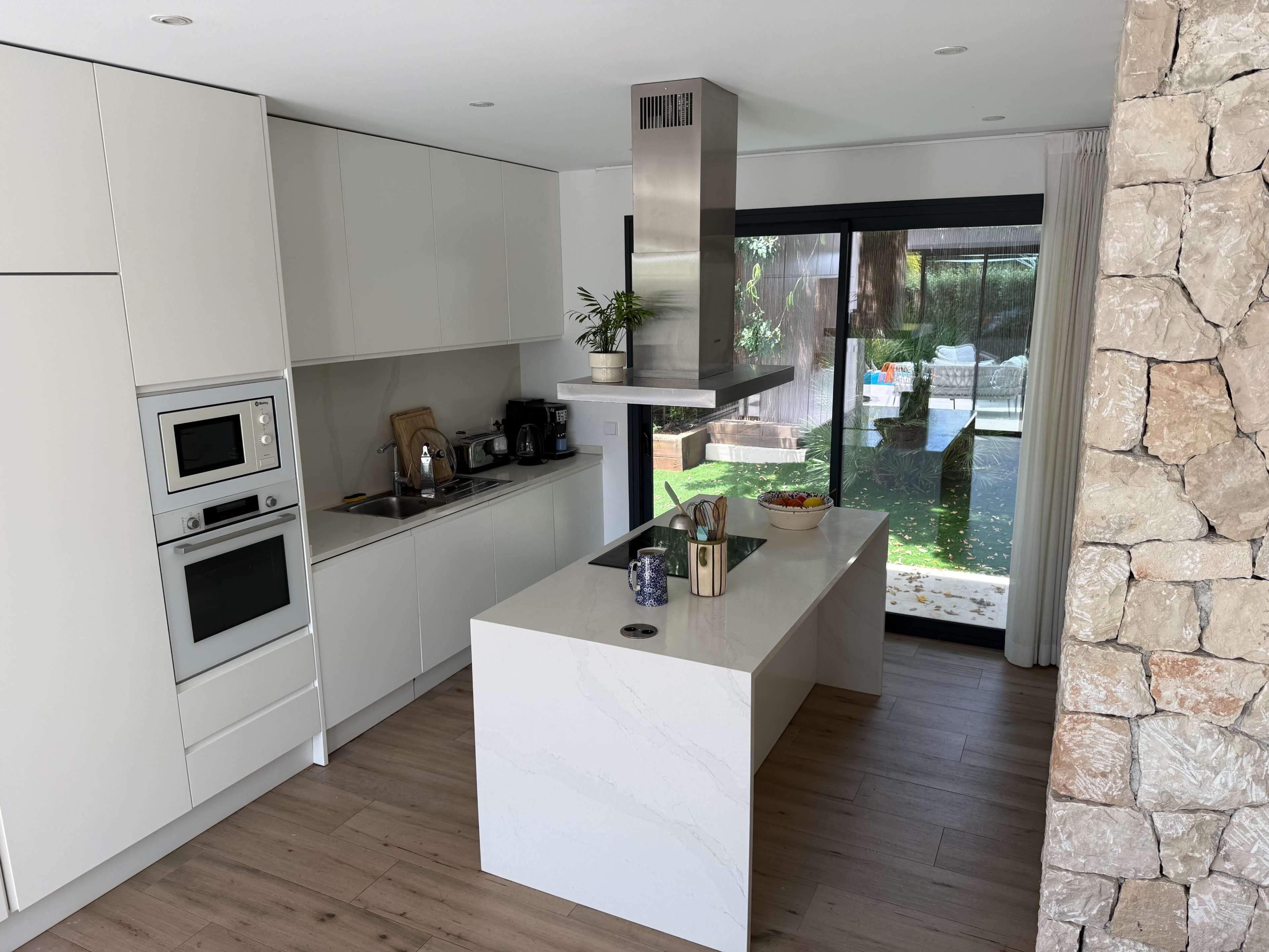 The image shows a modern kitchen with white cabinets, a central island, stainless steel appliances, and a large window overlooking a garden.