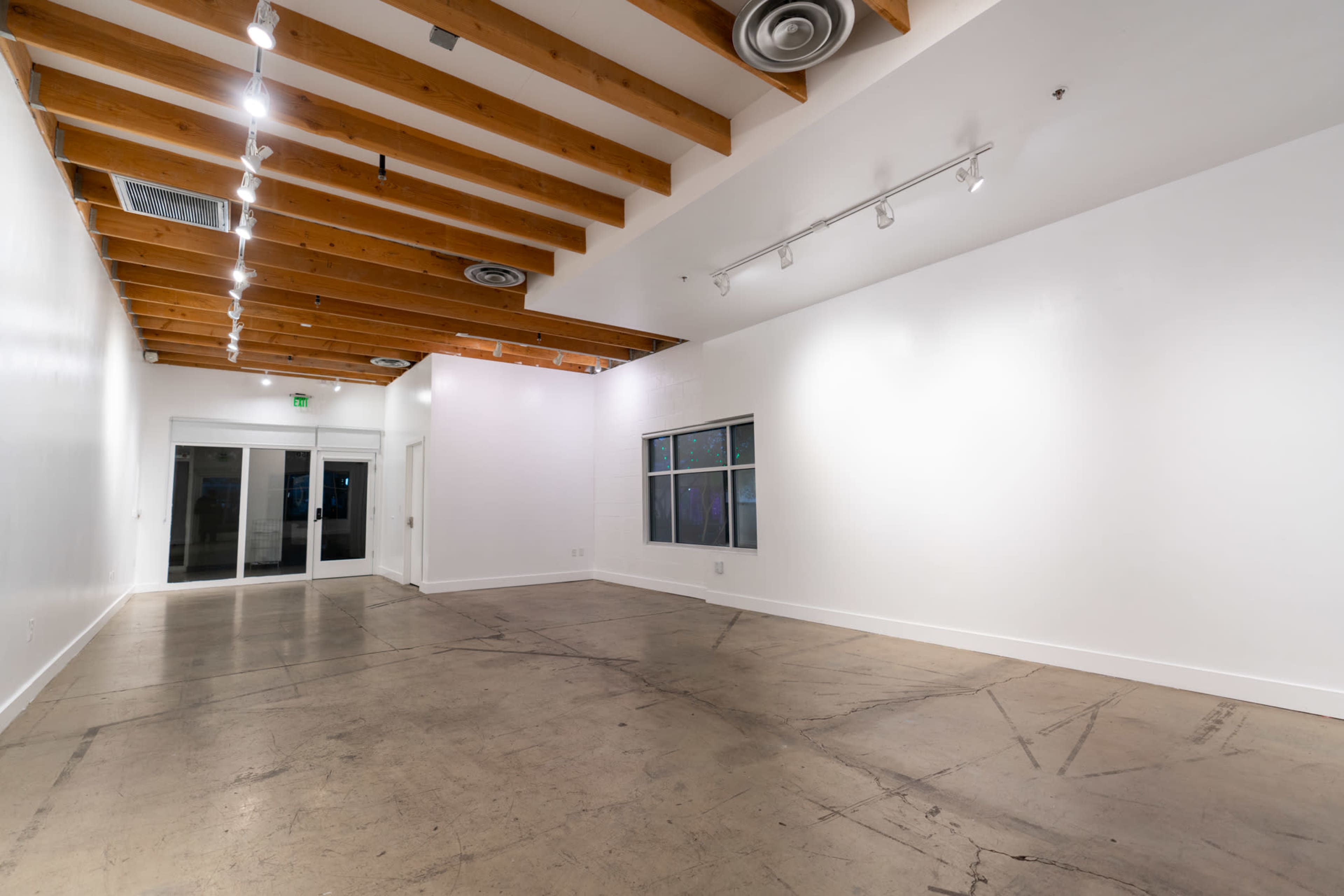 A vacant interior space with white walls, exposed wooden beams, and a concrete floor, featuring overhead lighting and large windows.