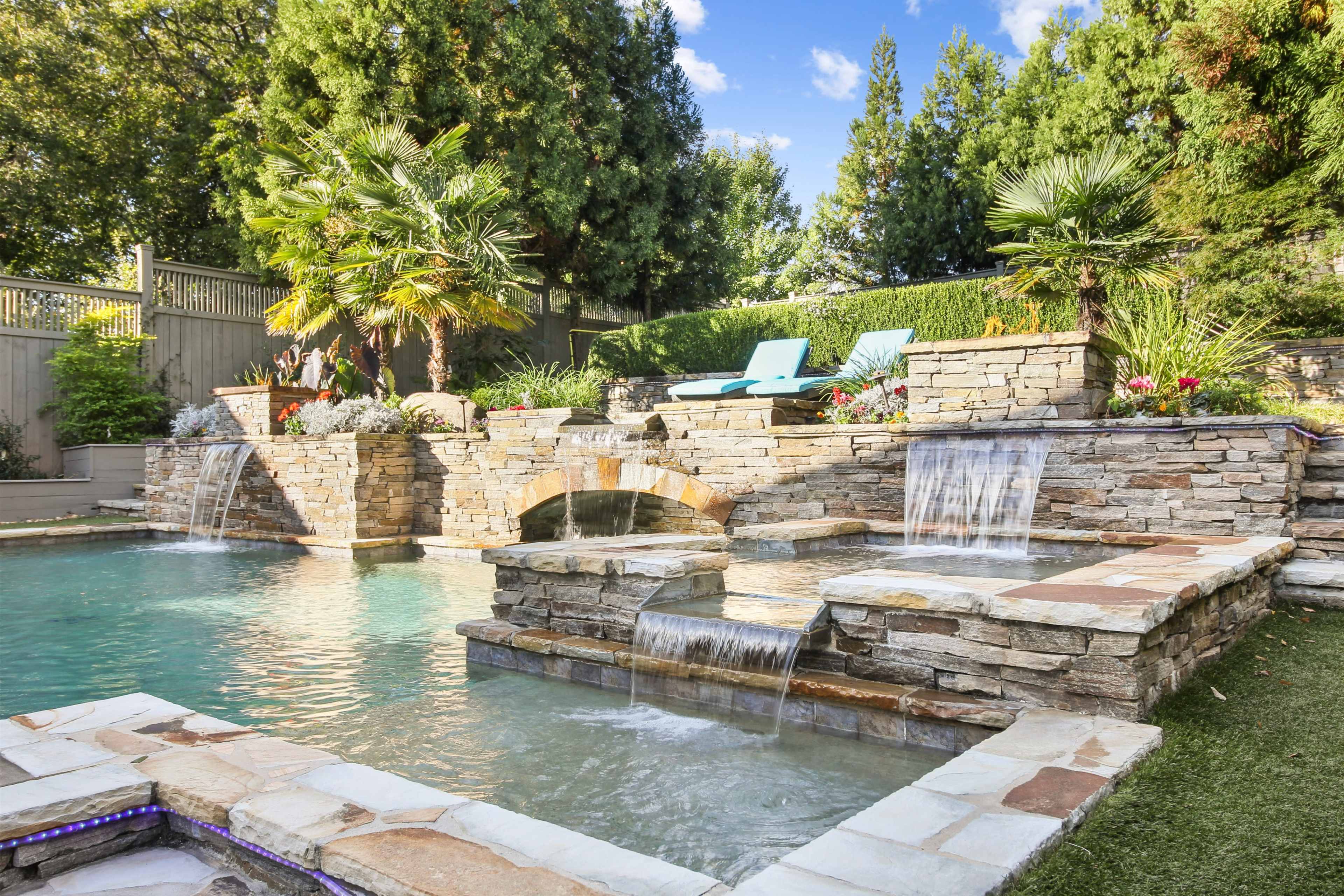 A landscaped pool area with stone waterfalls, a small bridge, and lounge chairs surrounded by lush greenery.