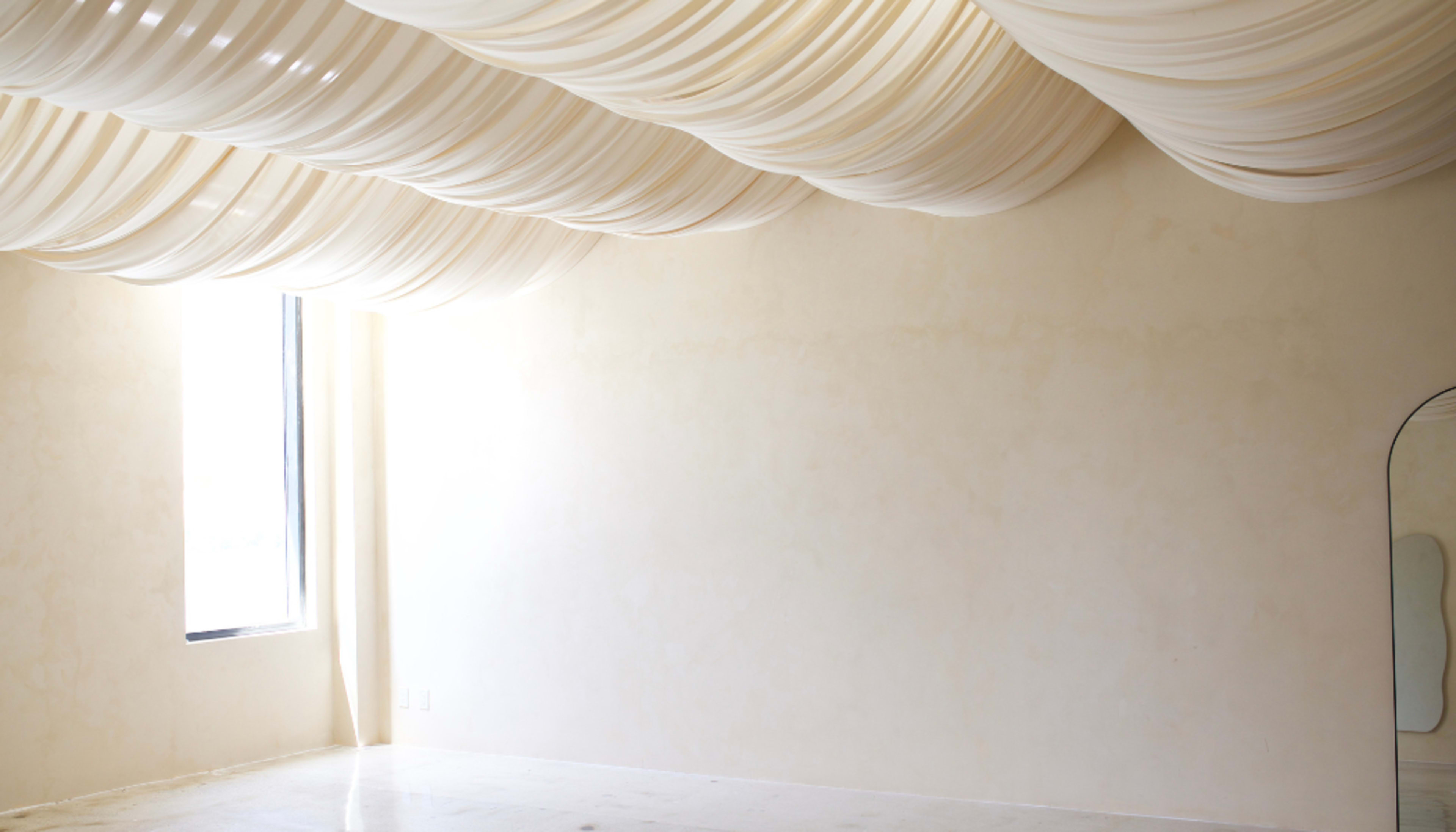 The image shows a spacious interior with soft, draped fabric across the ceiling and a large window allowing natural light to enter.