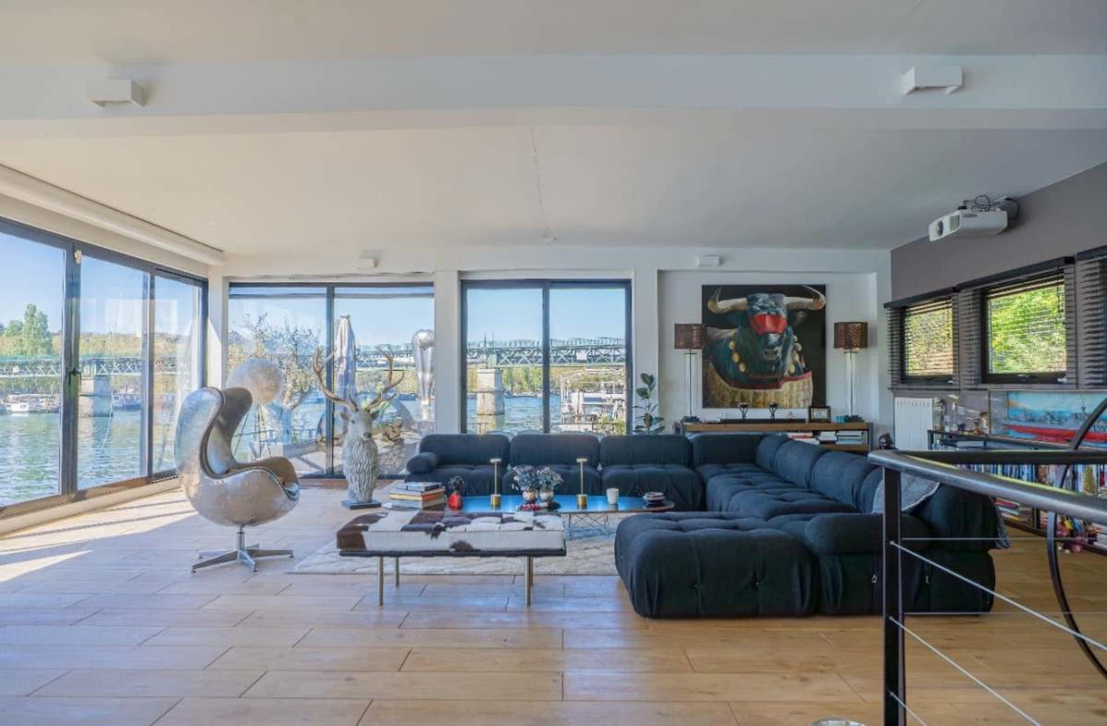 The image shows a spacious, modern living room with large windows overlooking a body of water and a bridge, featuring minimalist furniture and an abstract bull painting on the wall.