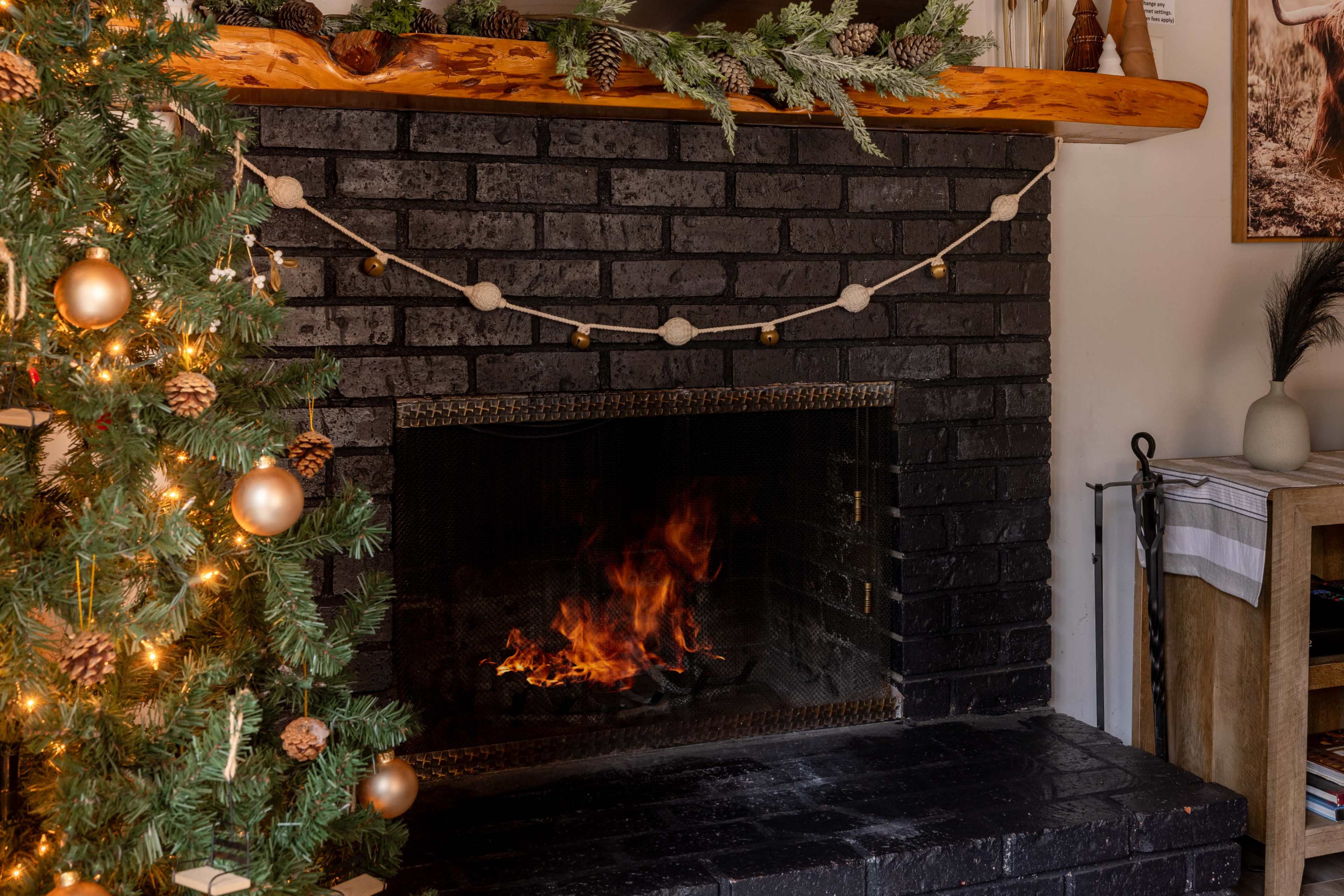 A cozy fireplace with a burning flame is framed by a black brick surround and decorated with a string of garland above, while a Christmas tree stands to the left adorned with ornaments.