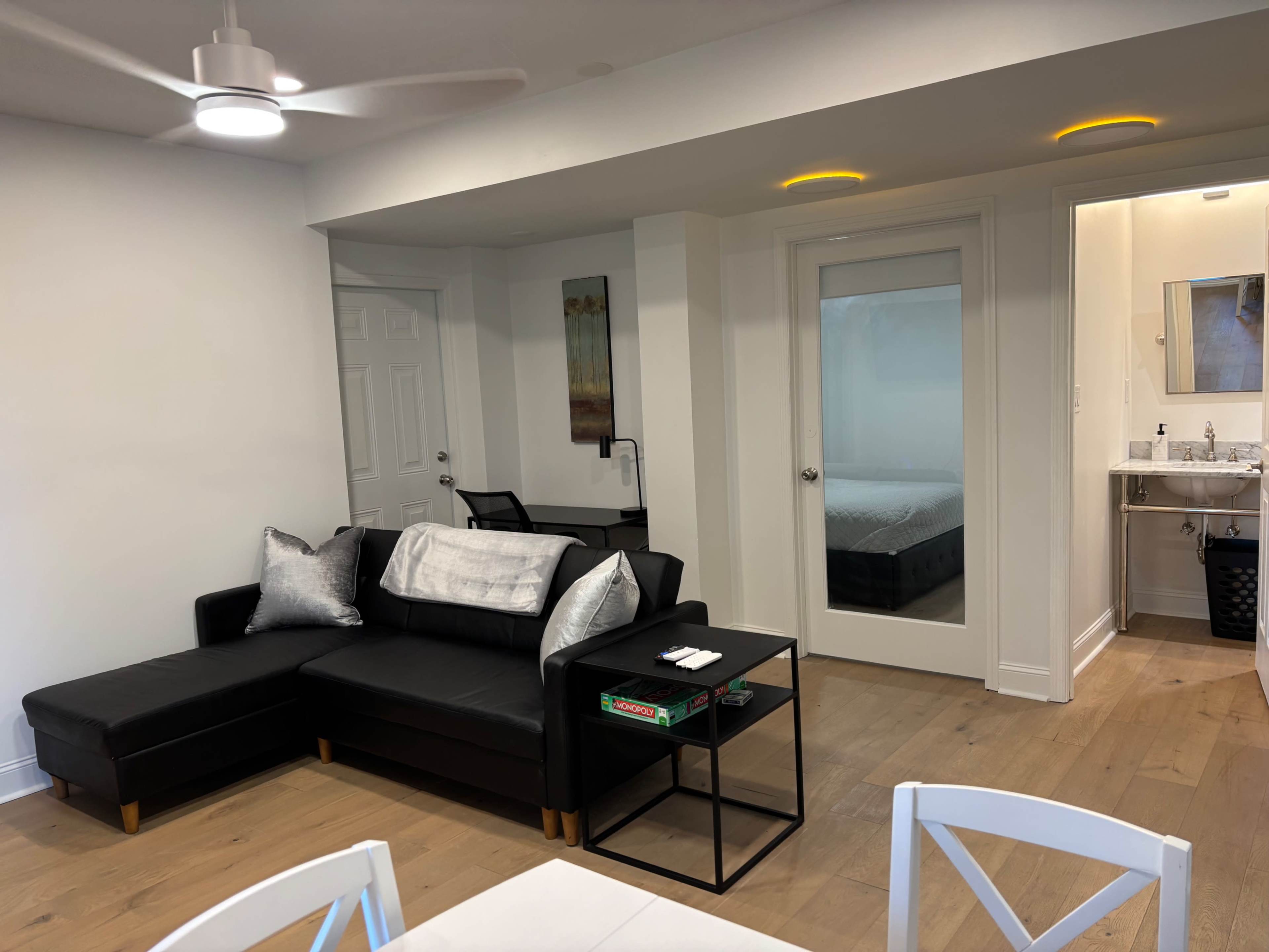 The image shows a contemporary living room with a black sectional sofa, a small glass coffee table, and a door leading to a bedroom and bathroom area.