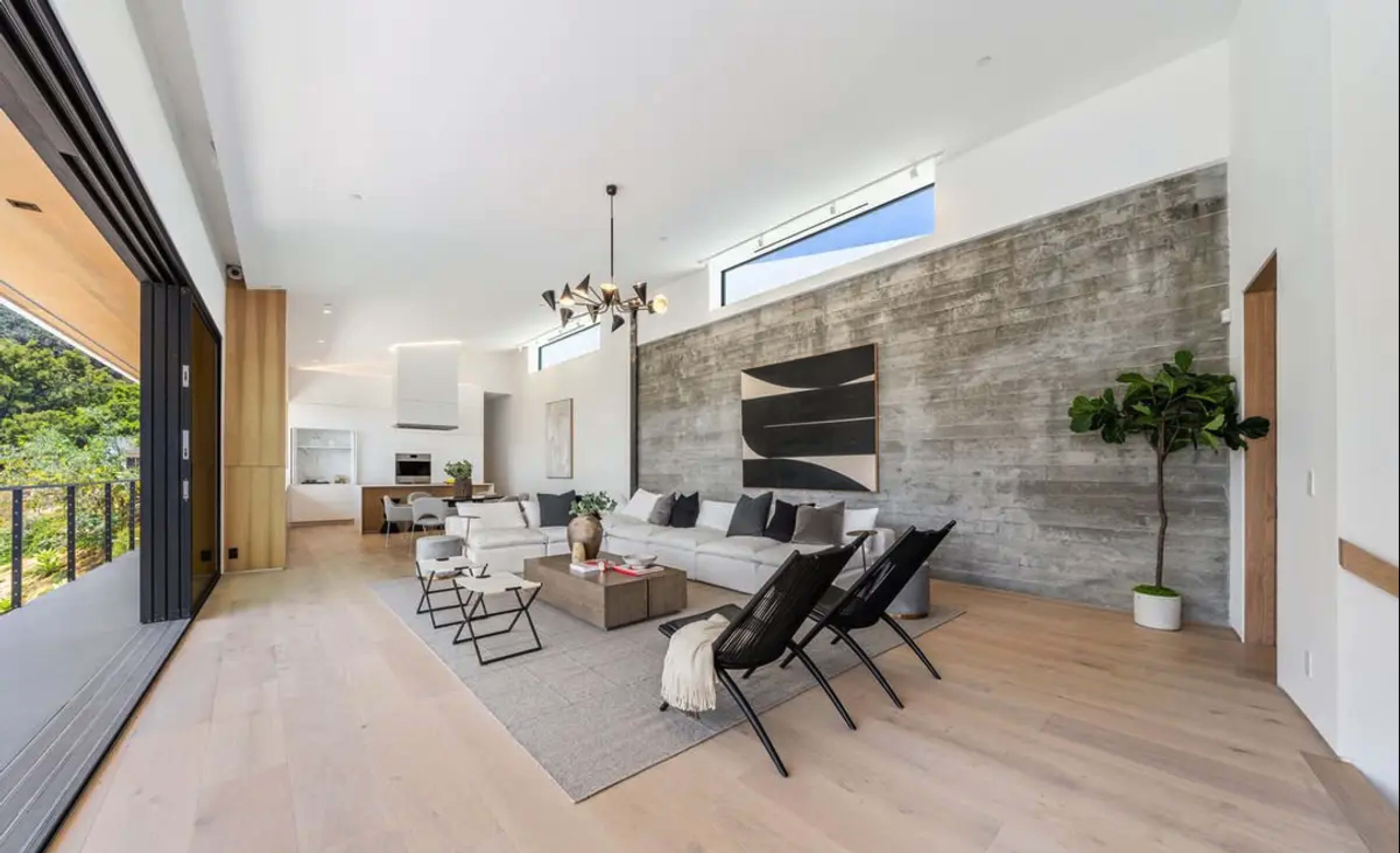 The image shows a modern living room featuring a large sectional sofa, minimalistic decor, and a wall of exposed concrete, with large windows providing natural light.