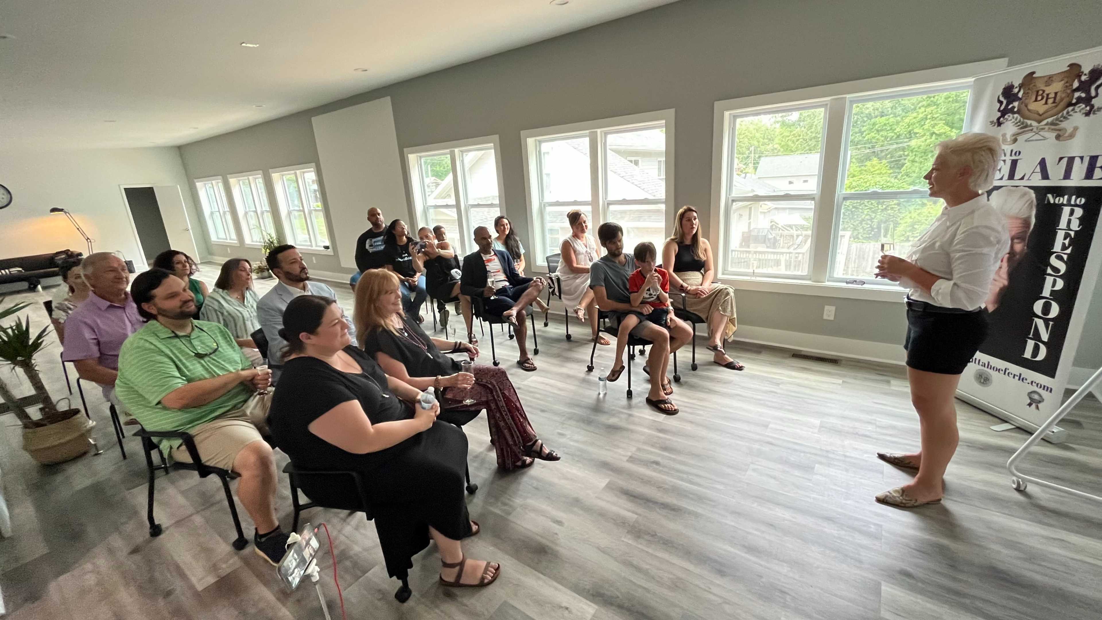 A group of people sits in a modern room with large windows while a woman stands at the front, speaking.