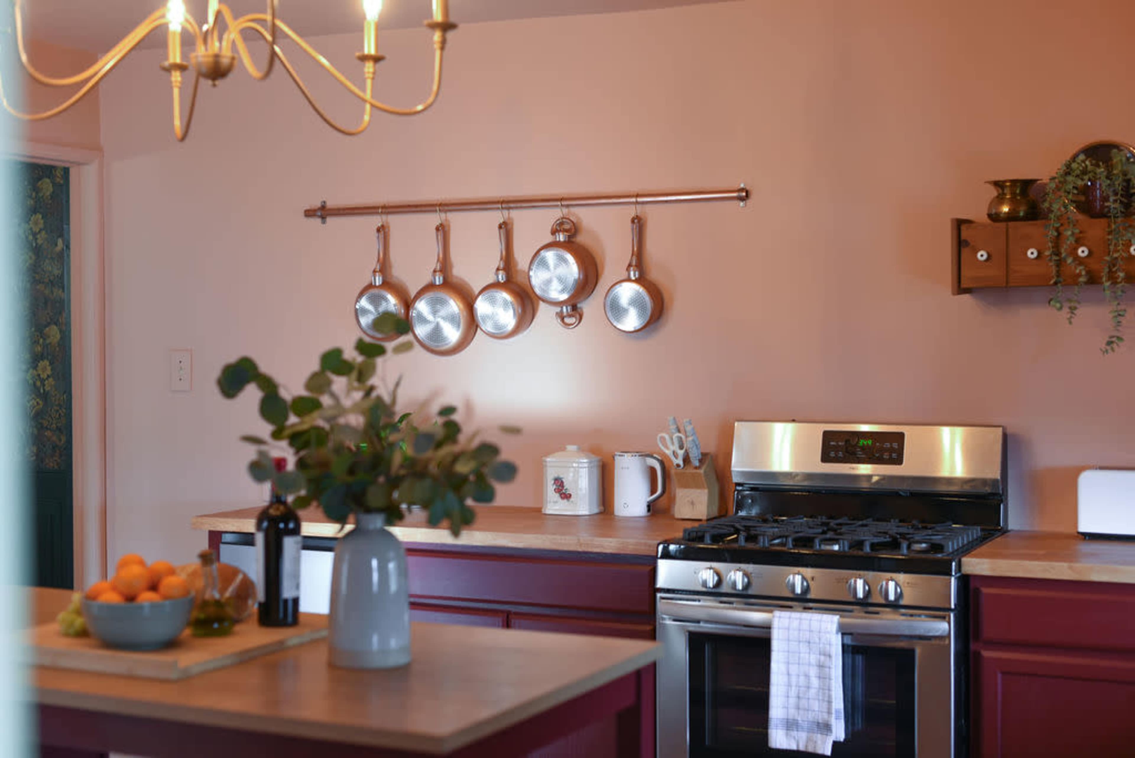 The image shows a kitchen with a gas stove, hanging copper pots, and a countertop featuring a vase of greenery and a bowl of oranges.