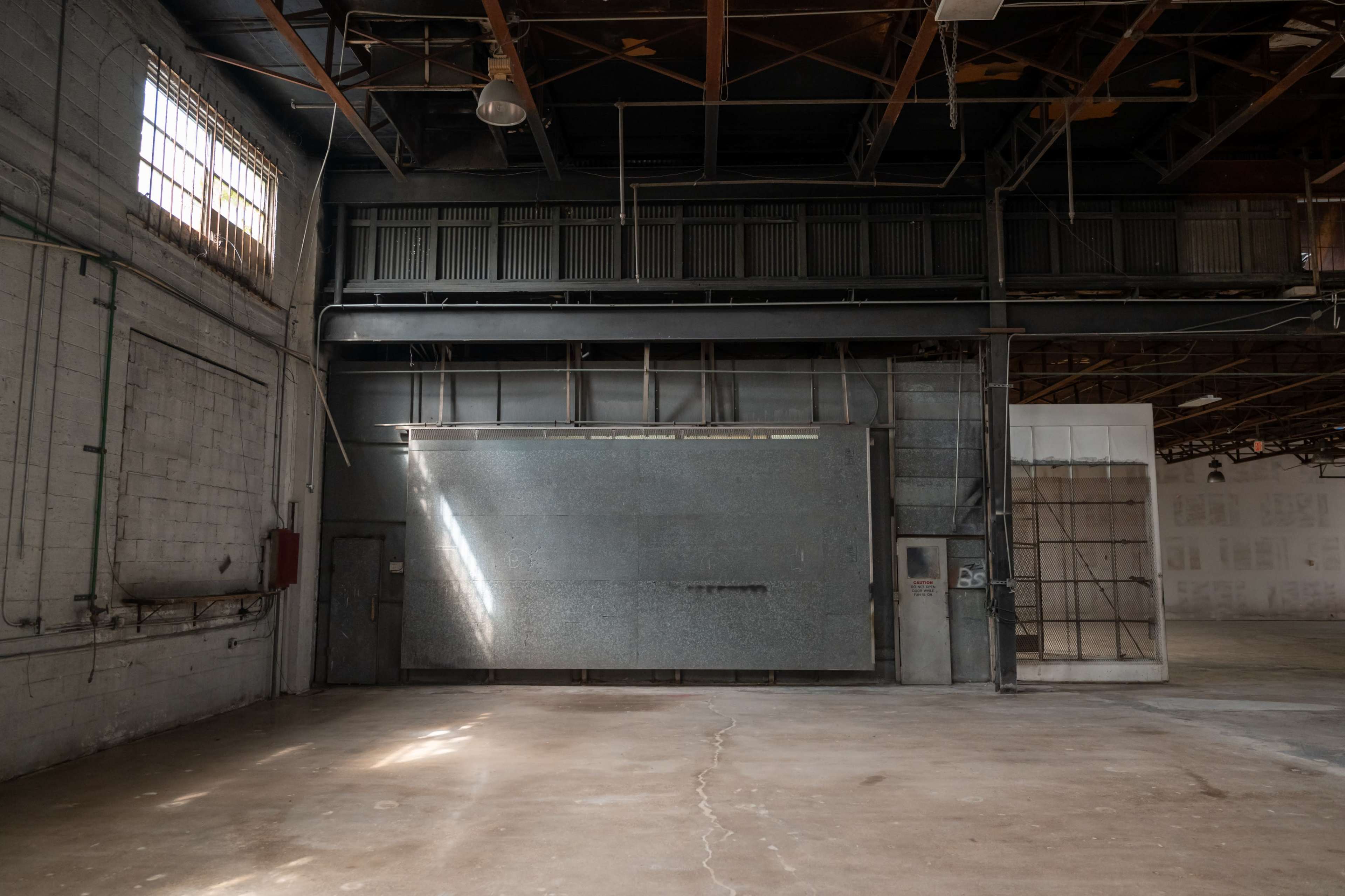 The image shows an empty industrial space with a large metal wall and a window, featuring exposed beams and concrete flooring.