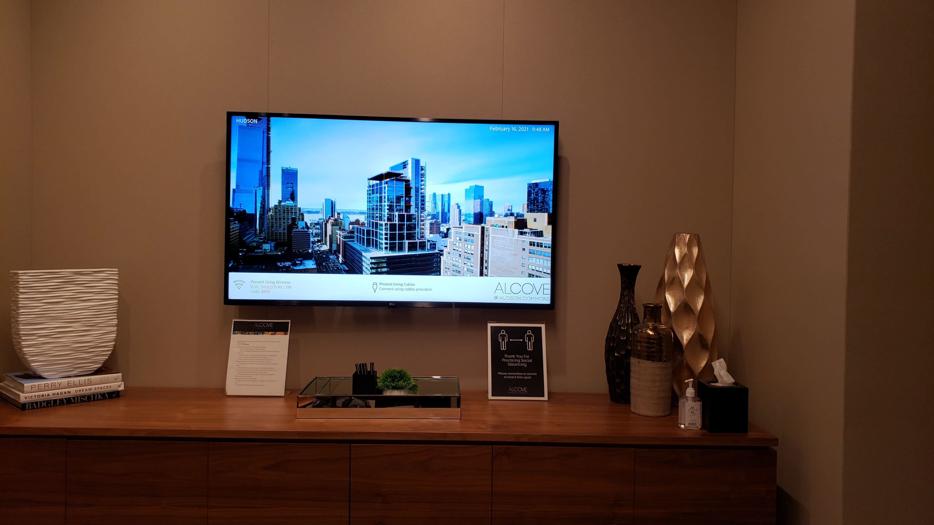 A modern room features a television displaying a cityscape, with a wooden console below that holds a plant, a tissue box, and decorative vases.