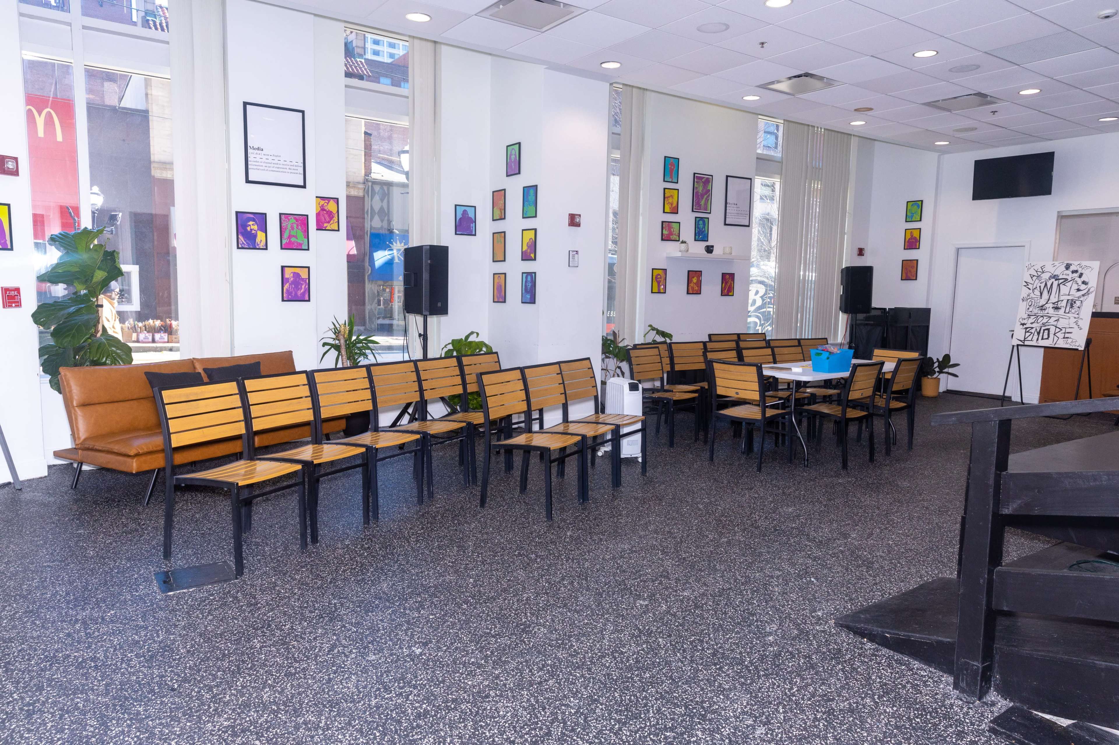 The image shows a modern interior space with rows of black metal chairs and a few tables arranged on a textured floor, alongside plants and colorful artwork on the walls.