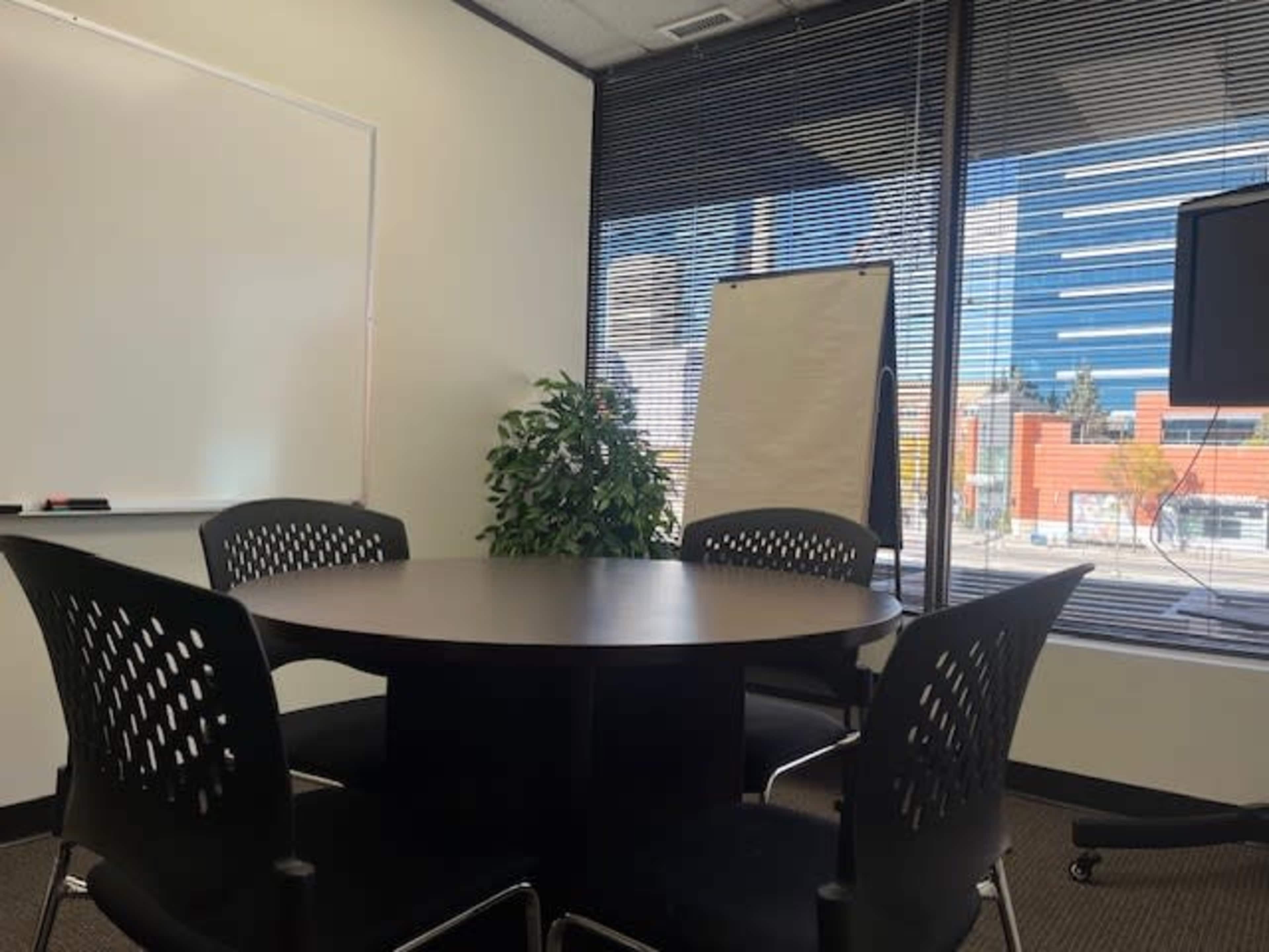 The image shows a small conference room with a round table and four black chairs, a whiteboard on the wall, and a large window revealing a view of a cityscape outside.