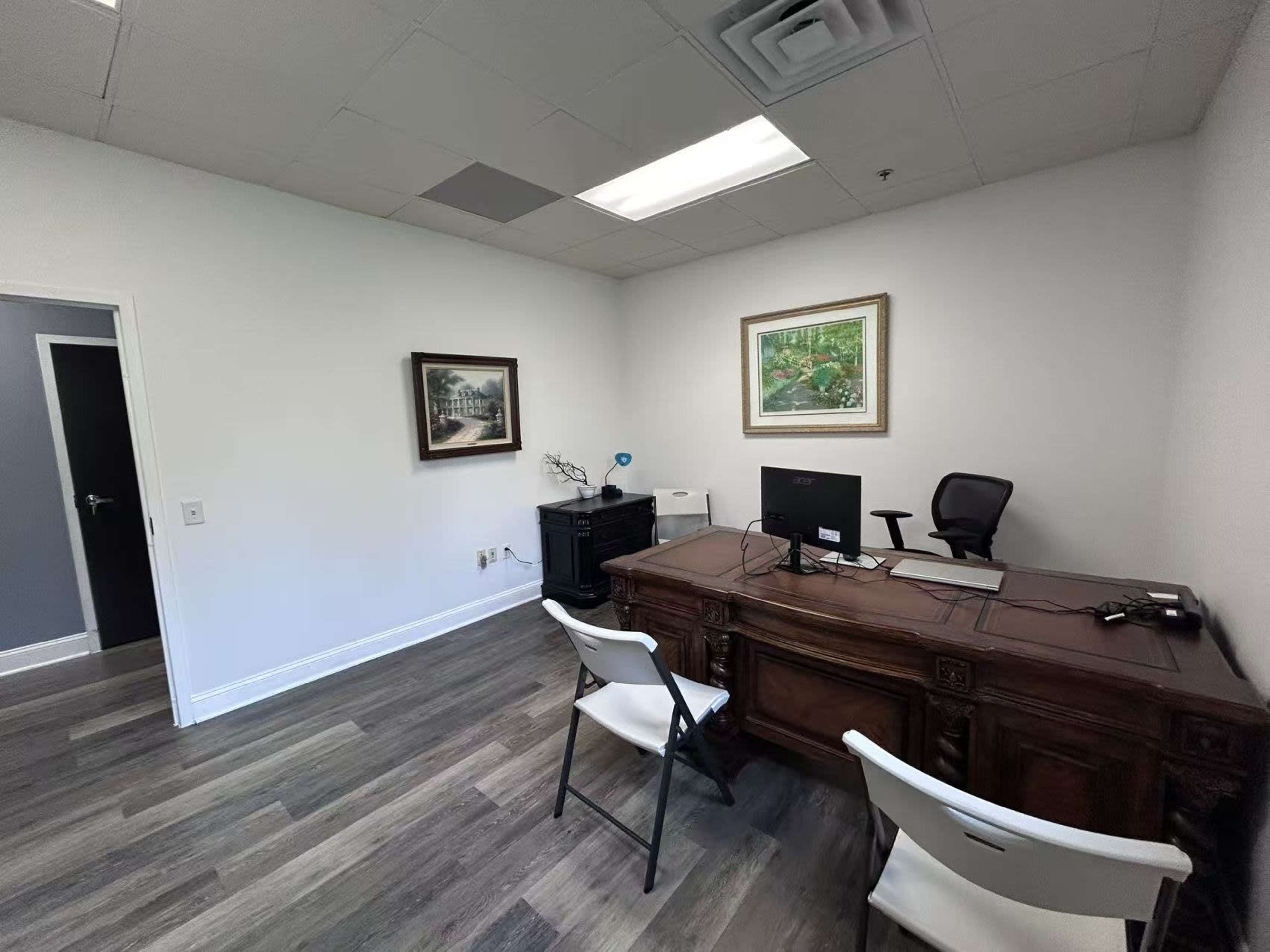 The image shows a sparsely furnished office with a large wooden desk, two chairs, and a computer setup against a plain wall, complemented by a framed painting and a small cabinet.