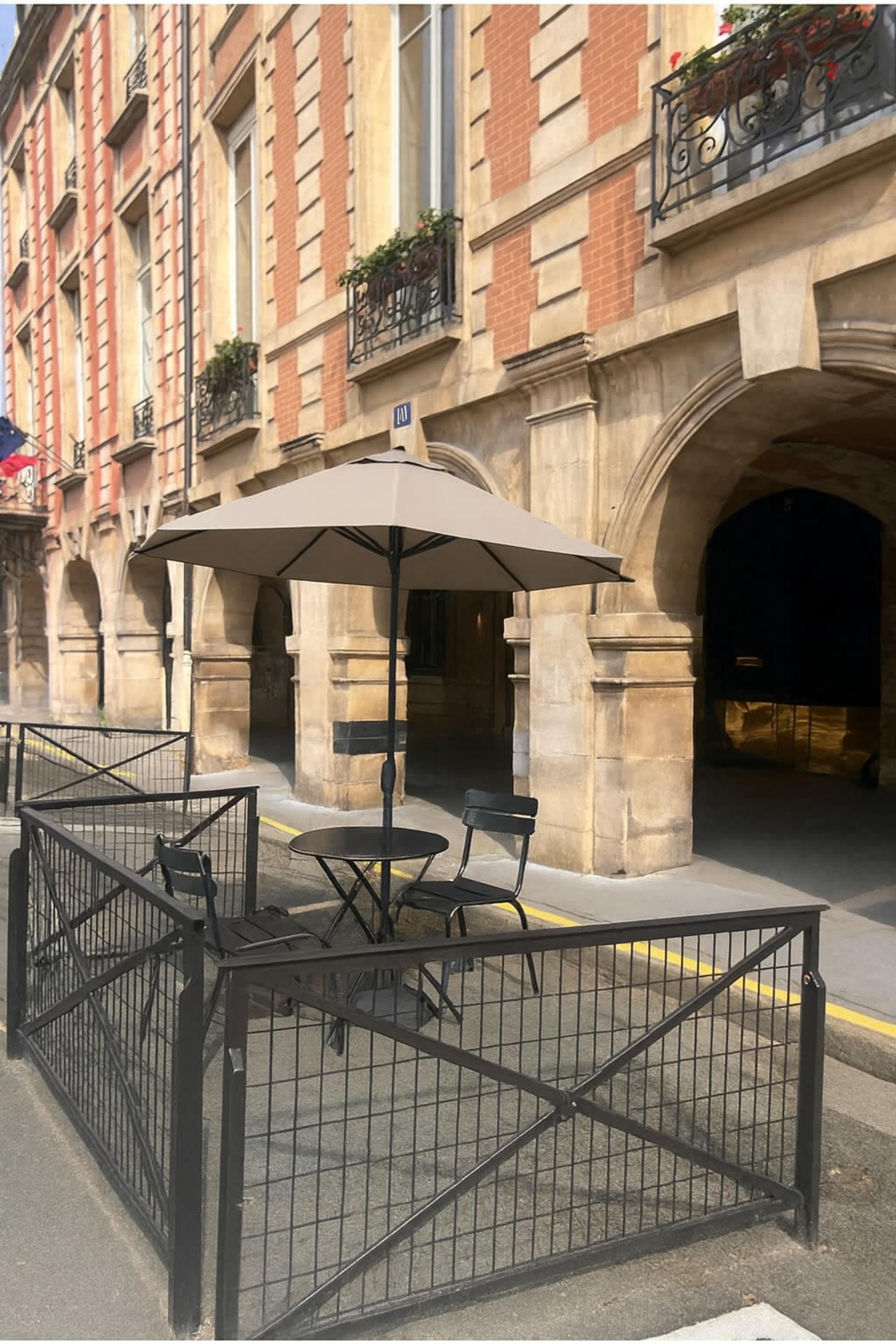 A small outdoor seating area with a circular table and two black chairs, sheltered by a large umbrella, is enclosed by a metal fence in front of a historic building.