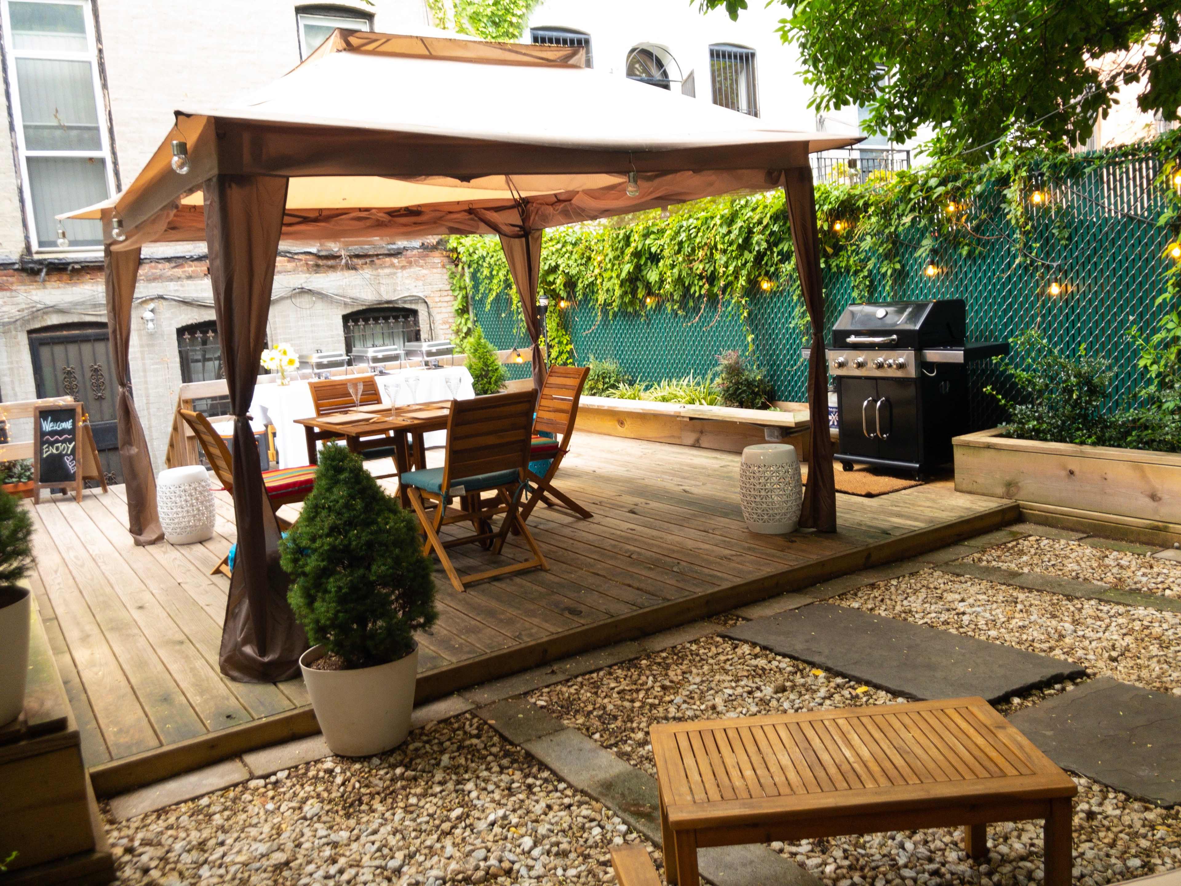 A wooden deck features a tented seating area with a dining table and chairs, accompanied by a barbecue grill nearby and surrounded by greenery.