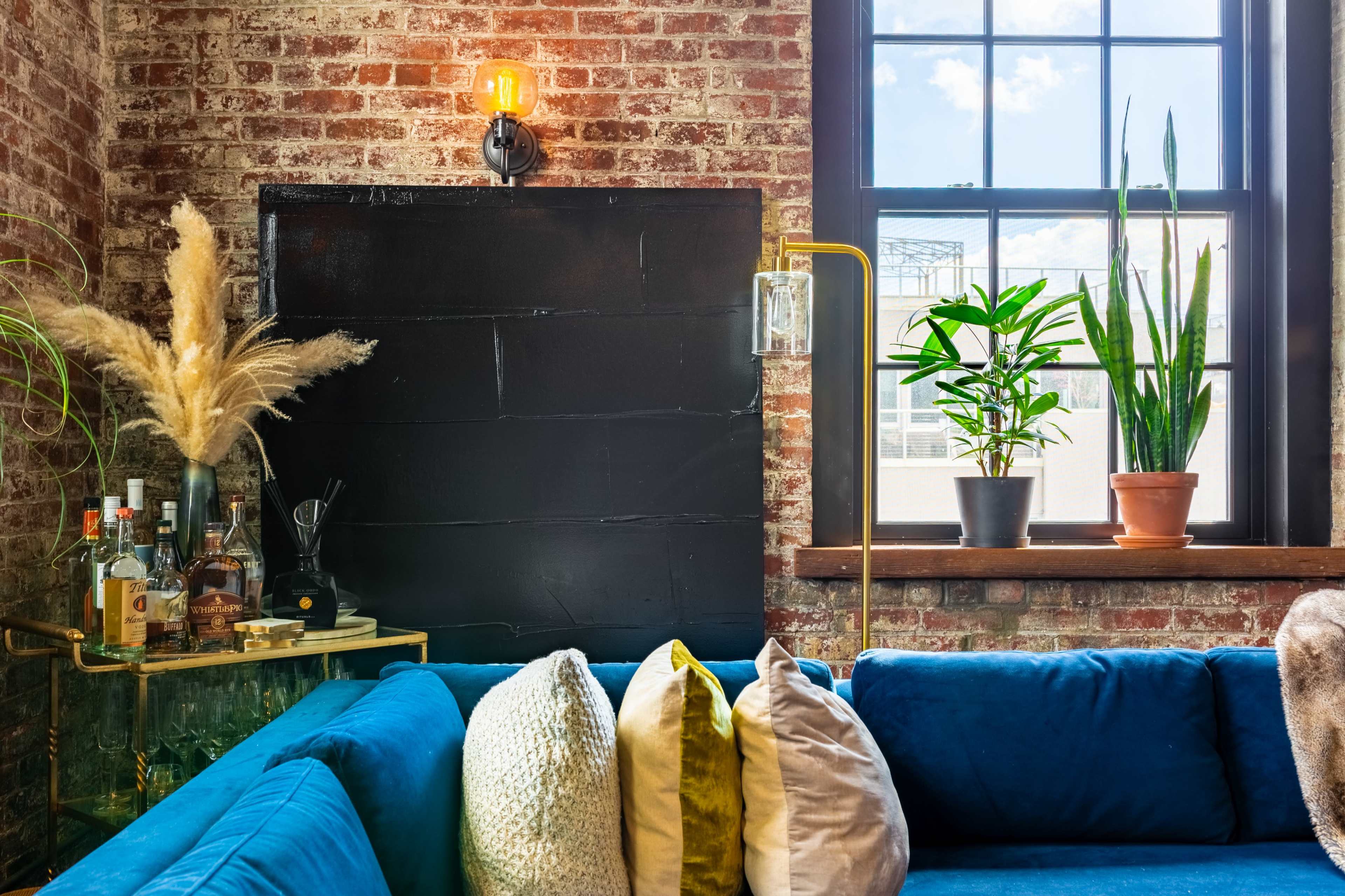 A blue sofa with decorative pillows is positioned near a window, showcasing plants and a small bar cart against a brick wall.