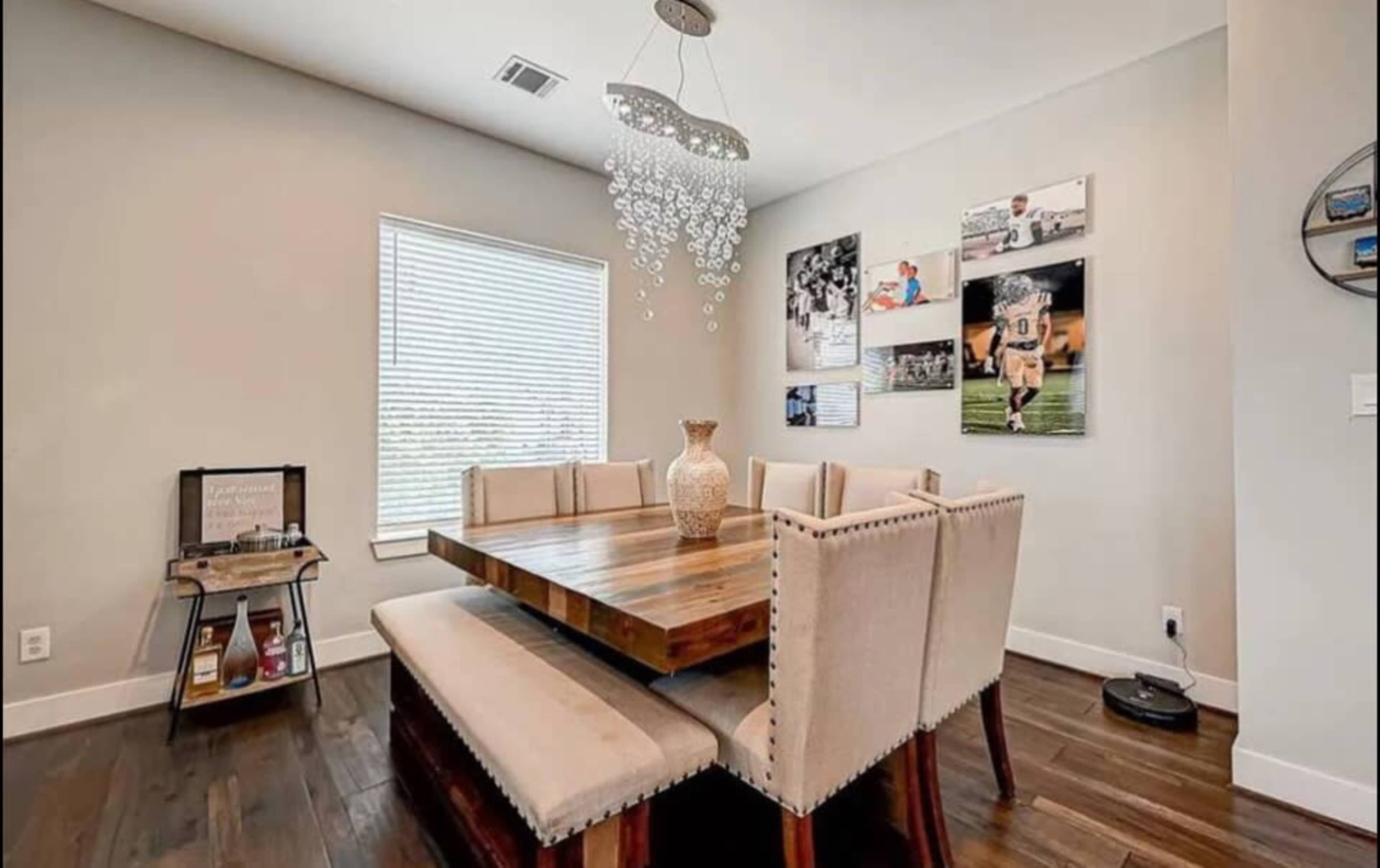 A dining area features a wooden table surrounded by upholstered benches, illuminated by a modern chandelier, with framed photographs on the walls.