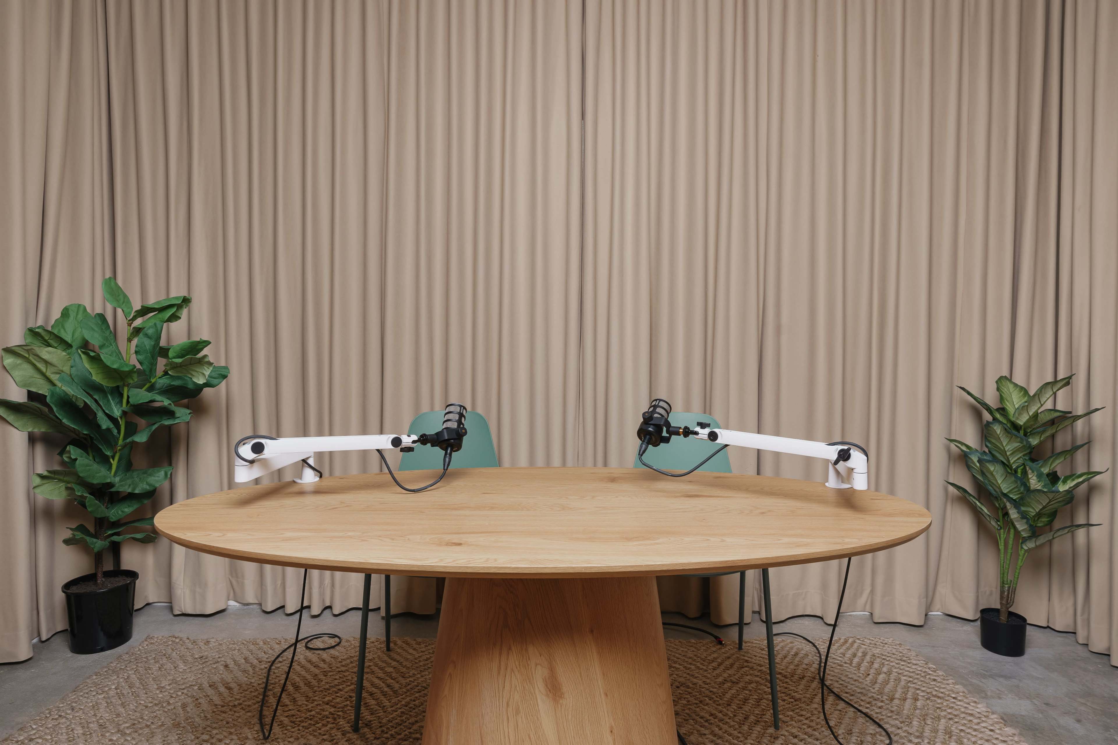 A podcast studio setup with a large round table, two chairs, and microphone stands against a backdrop of beige curtains and potted plants.