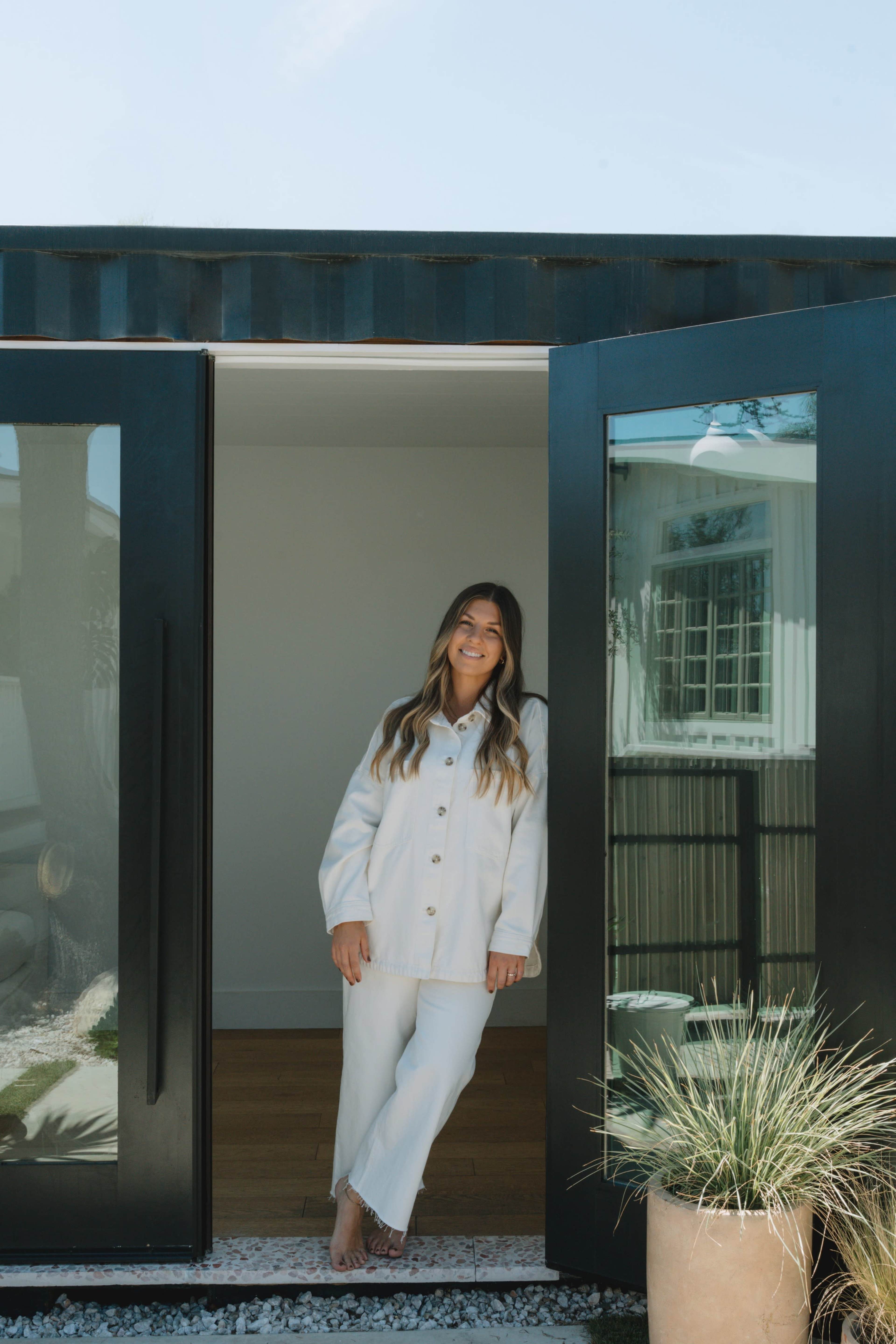 A woman stands smiling in the doorway of a modern, minimalist building surrounded by greenery.