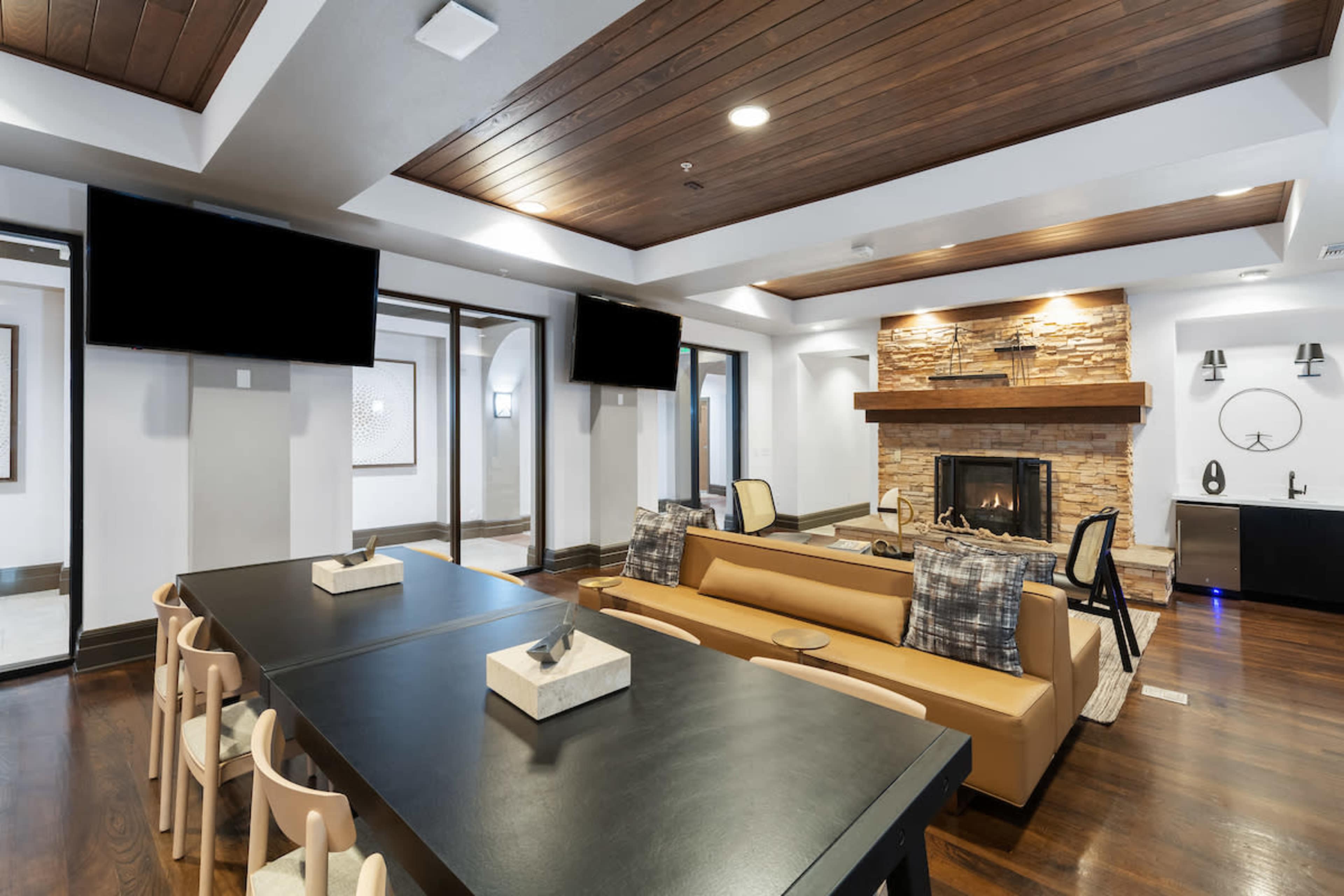 A modern lounge area with a wooden ceiling, a stone fireplace, a long dining table, comfortable seating, and wall-mounted televisions.