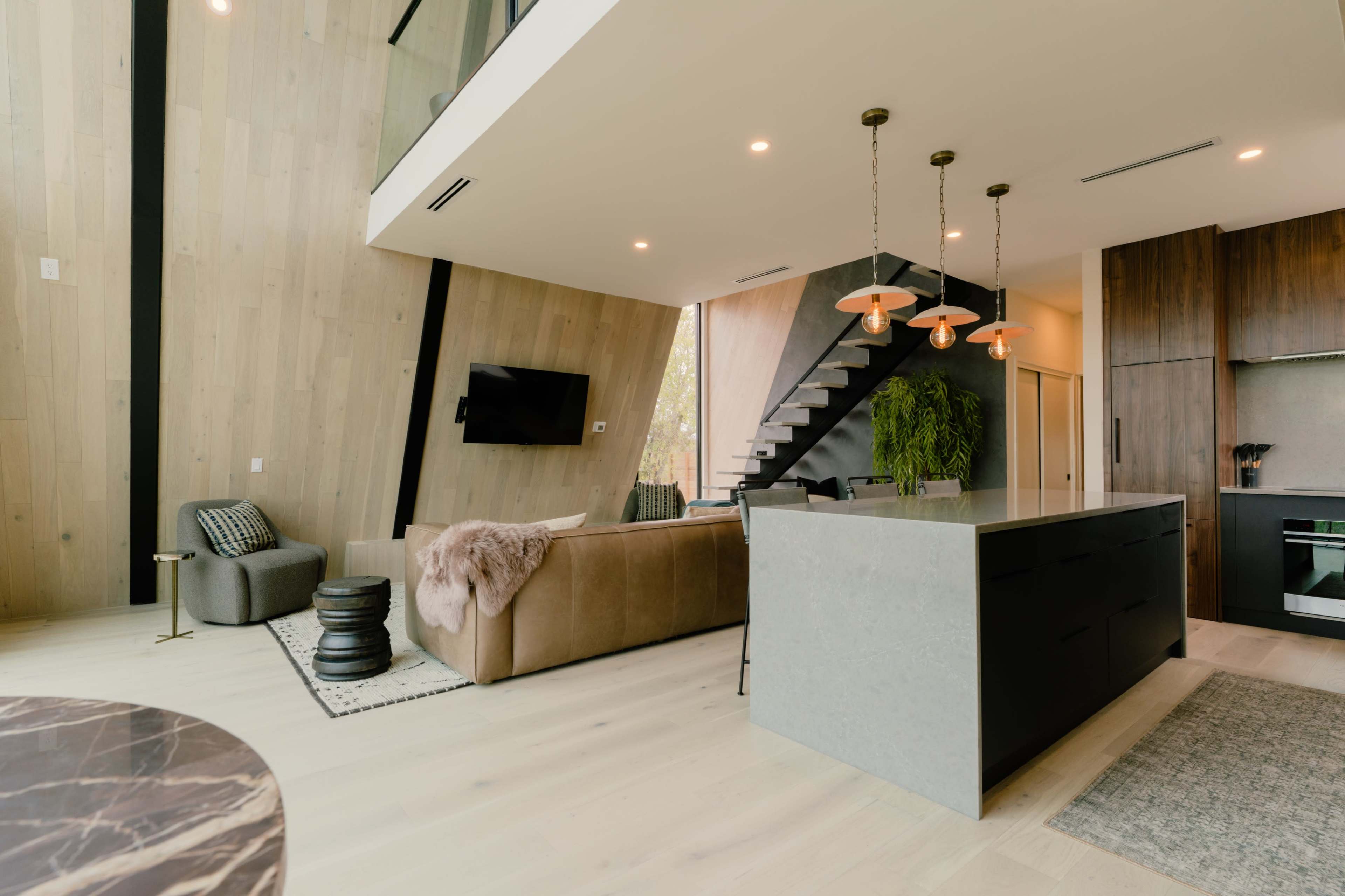 A modern living space features a beige sofa, a marble-topped dining table, a kitchen island, and a staircase leading to an upper level.