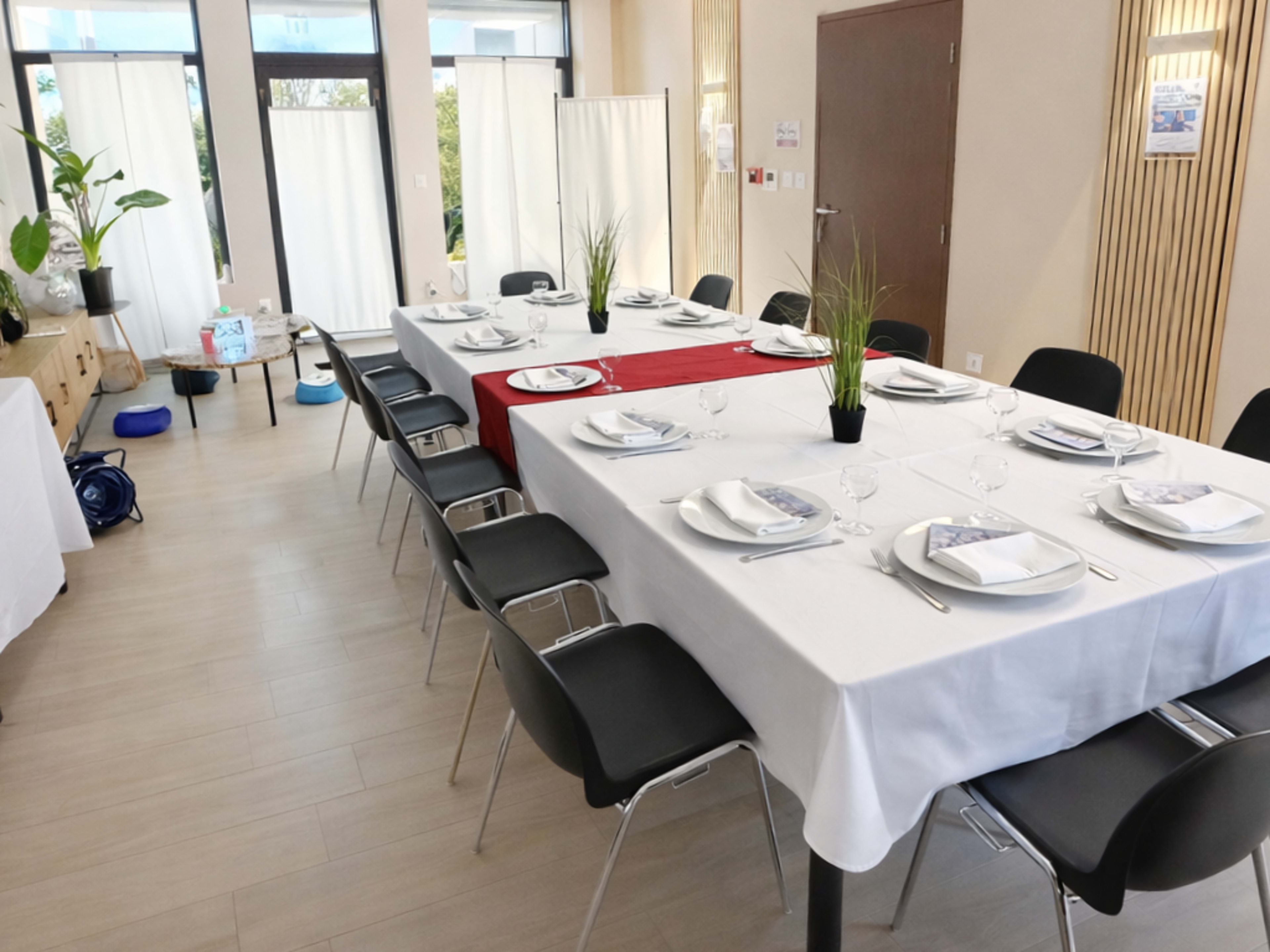 A dining table is set with white tablecloths and black chairs, featuring plates, glasses, and decorative plants, in a well-lit room.