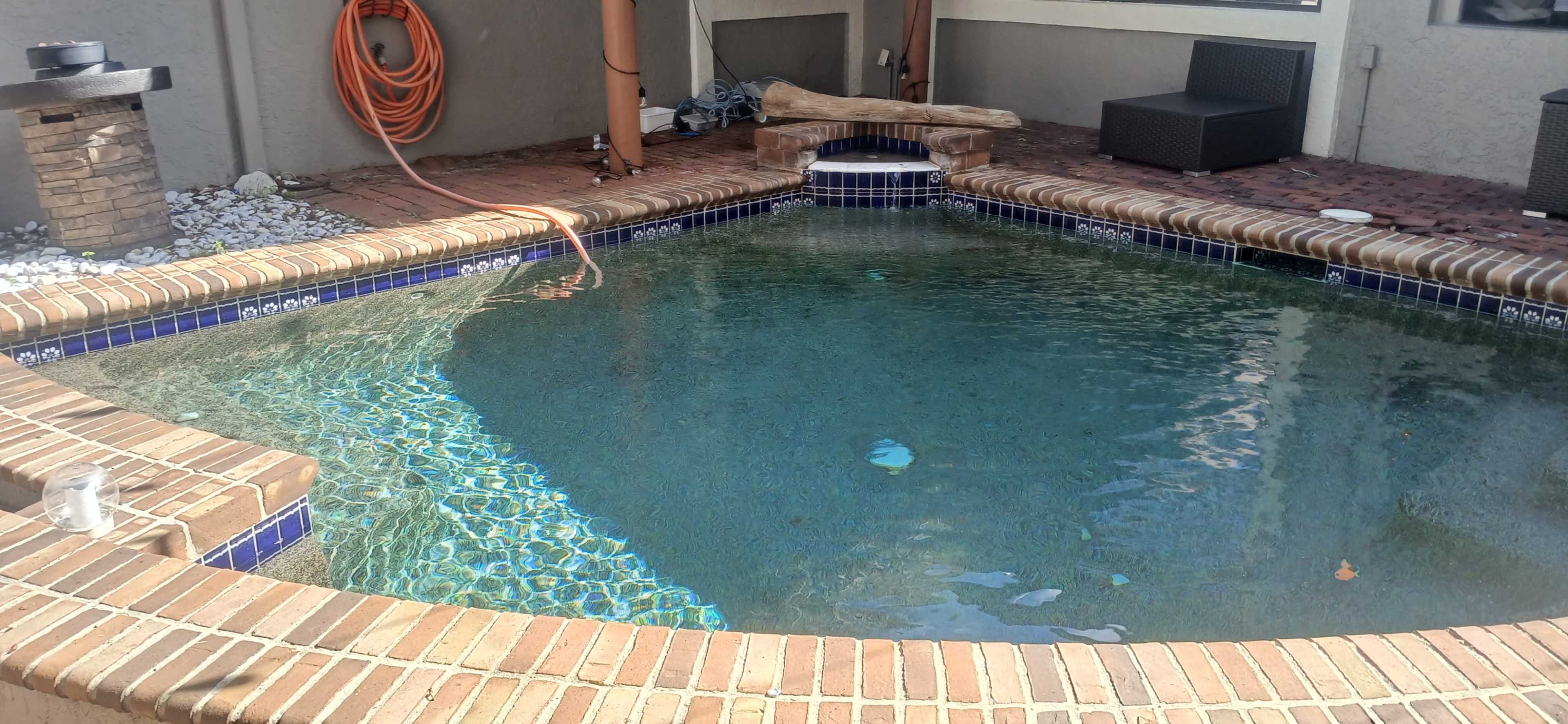 The image shows a rectangular swimming pool with clear water, surrounded by brick and stone walls, and featuring outdoor furniture nearby.