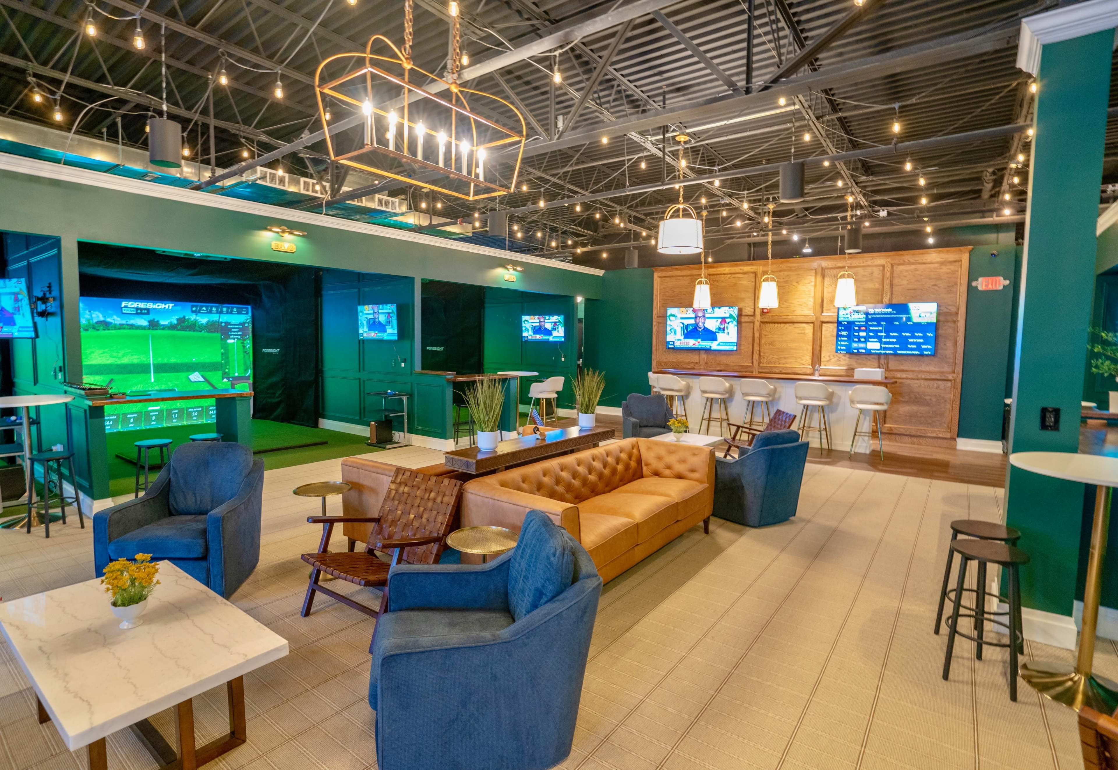 The image shows a modern indoor lounge with comfortable seating, multiple televisions displaying sports, and a putting green area in the background.