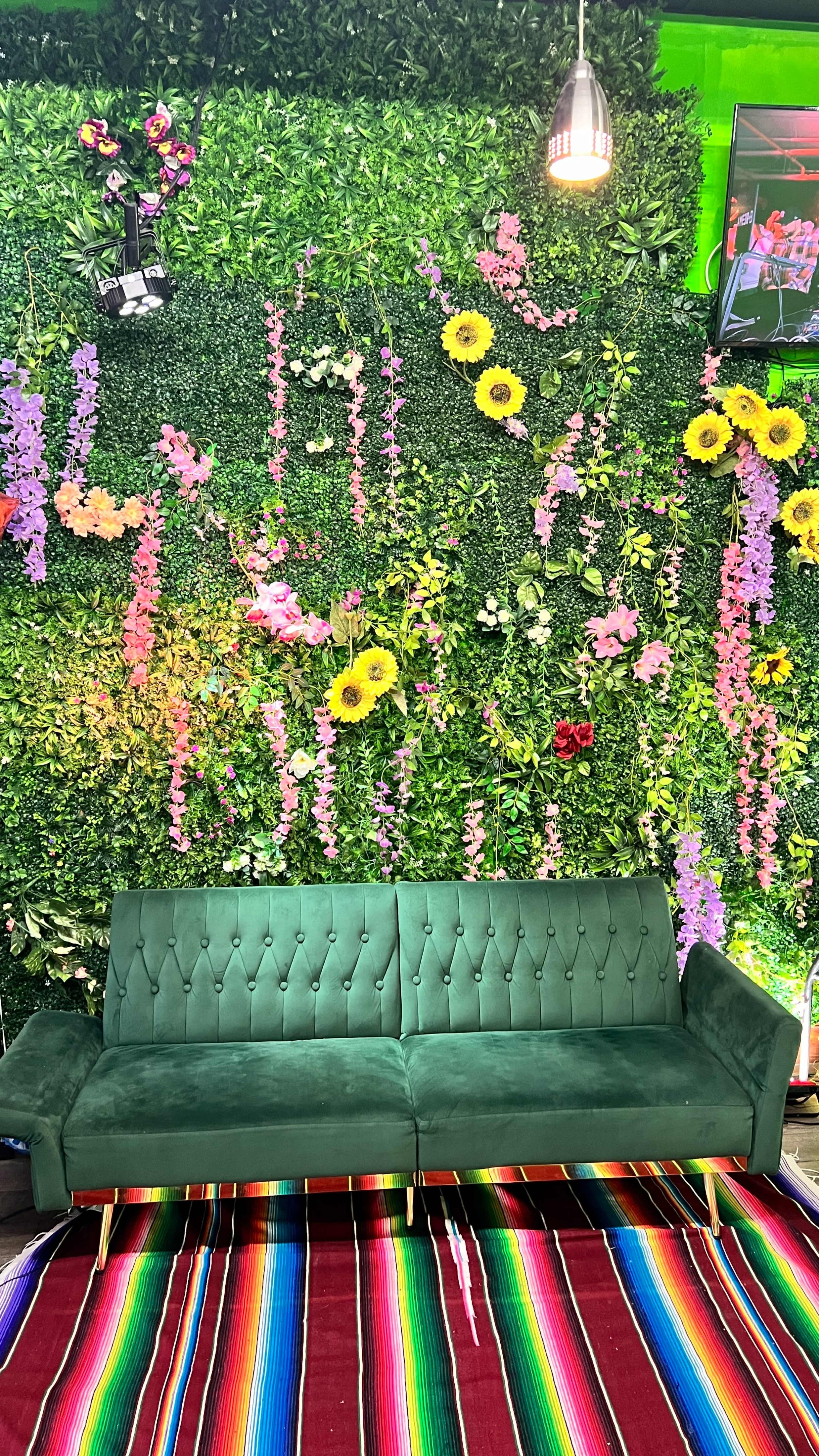 The image features a green velvet couch in front of a wall adorned with various artificial flowers and plants, along with a colorful striped rug underneath.