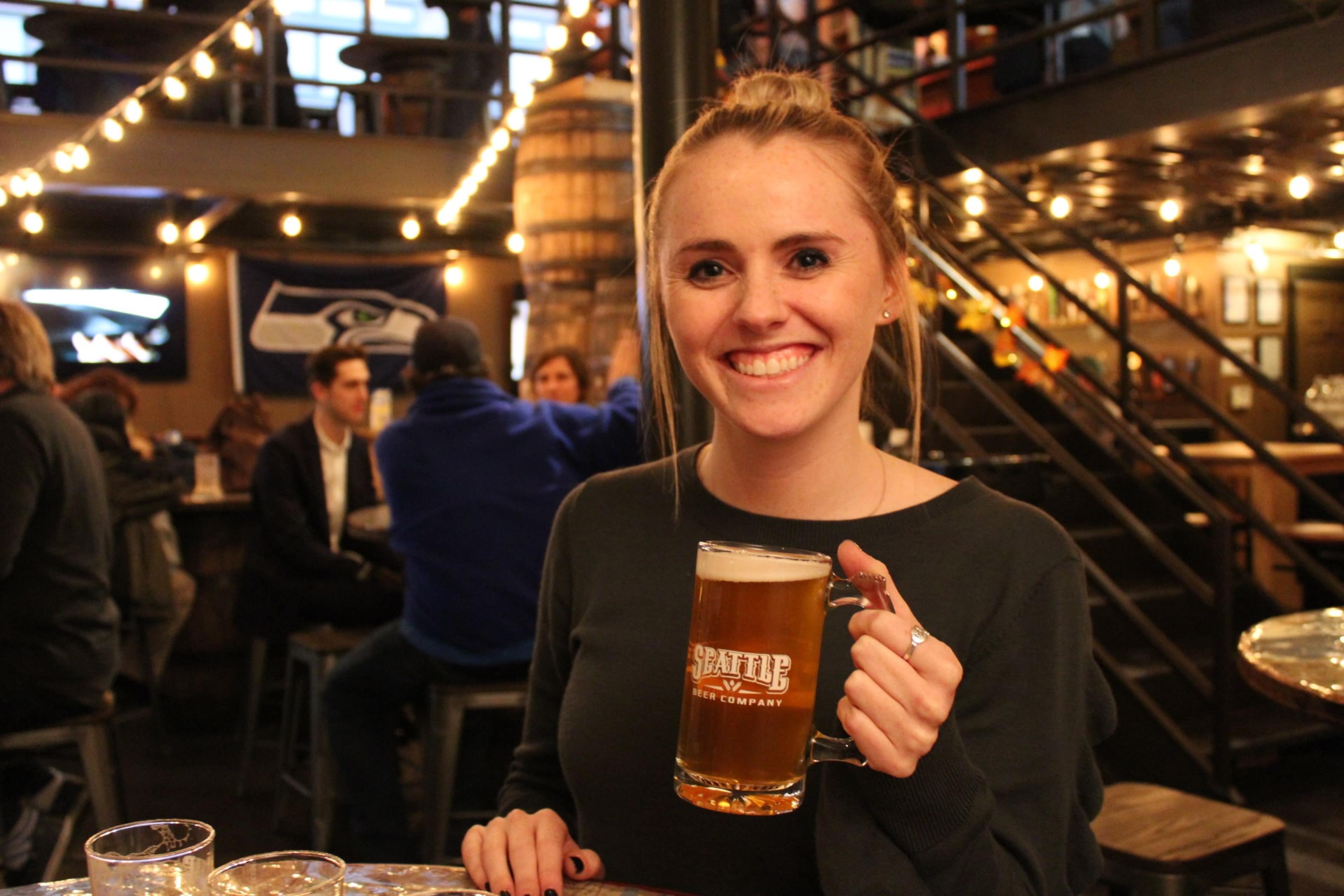 A woman smiles while holding a mug of beer in a bustling brewery with patrons in the background.